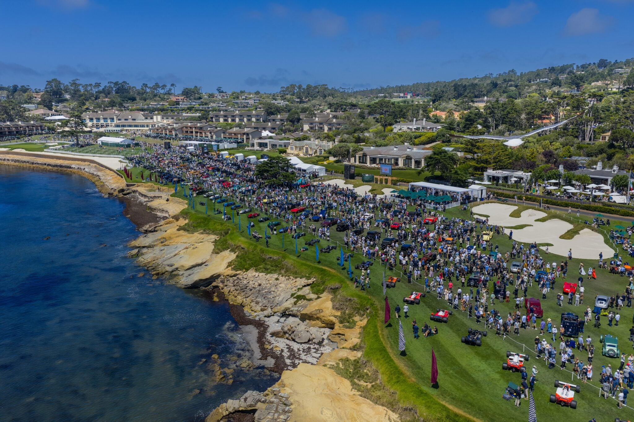 A large crowd gathers on a grassy area by the ocean for the Pebble Beach Concours d’Elegance, celebrating 75 years of automotive artistry, with classic cars on display and buildings in the background under a blue sky.
