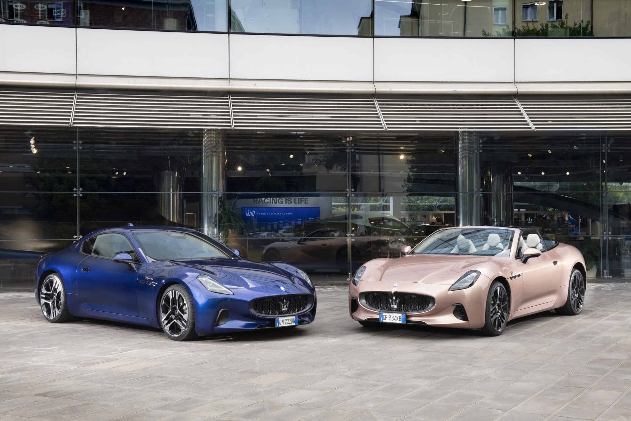 A blue Maserati GranTurismo coupe and a pink convertible sports car are parked side by side in front of a modern glass building in Modena.