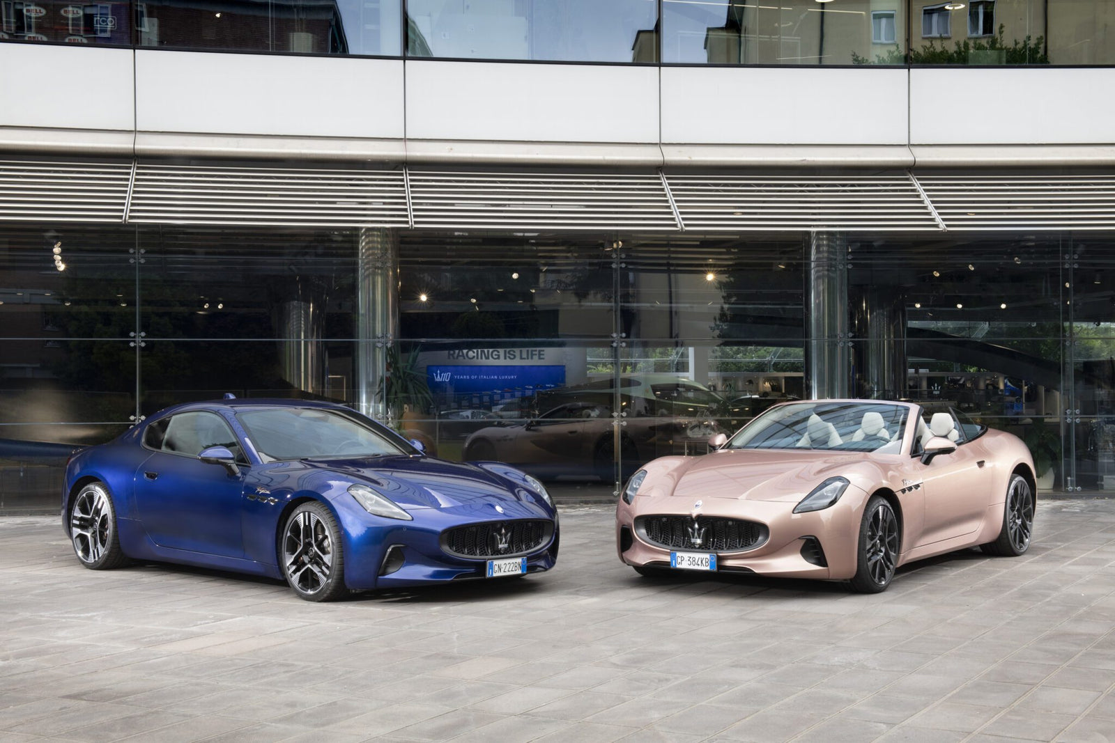 A blue Maserati GranTurismo coupe and a pink convertible sports car are parked side by side in front of a modern glass building in Modena.
