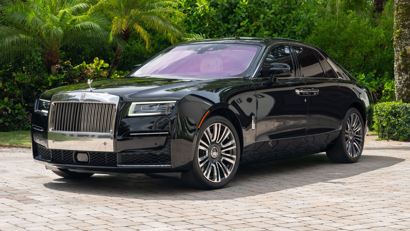 A 2024 Rolls-Royce Ghost collector’s car sedan parked on a stone driveway, surrounded by green foliage and palm trees.