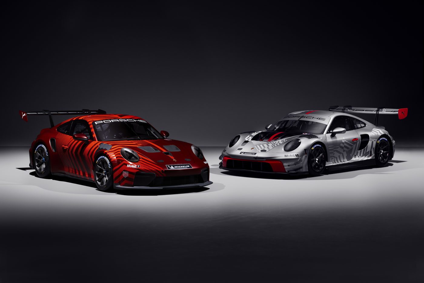 Two Porsche race cars—a red 911 GT3 R and a silver 911 Cup Car—are parked side by side in a dark, studio-lit setting.