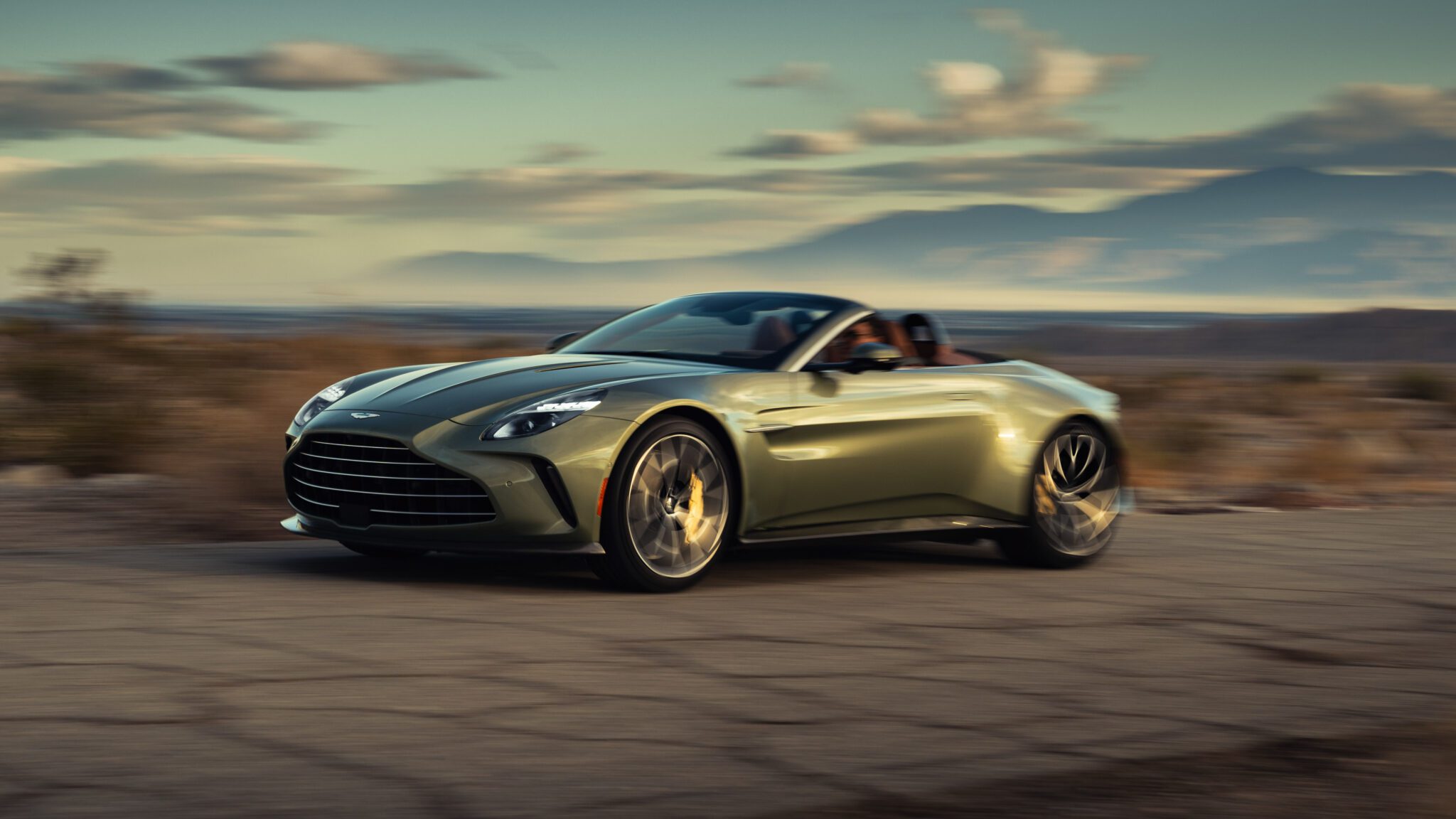 A green convertible sports car driving on a deserted road through a scenic, arid landscape with mountains in the background.