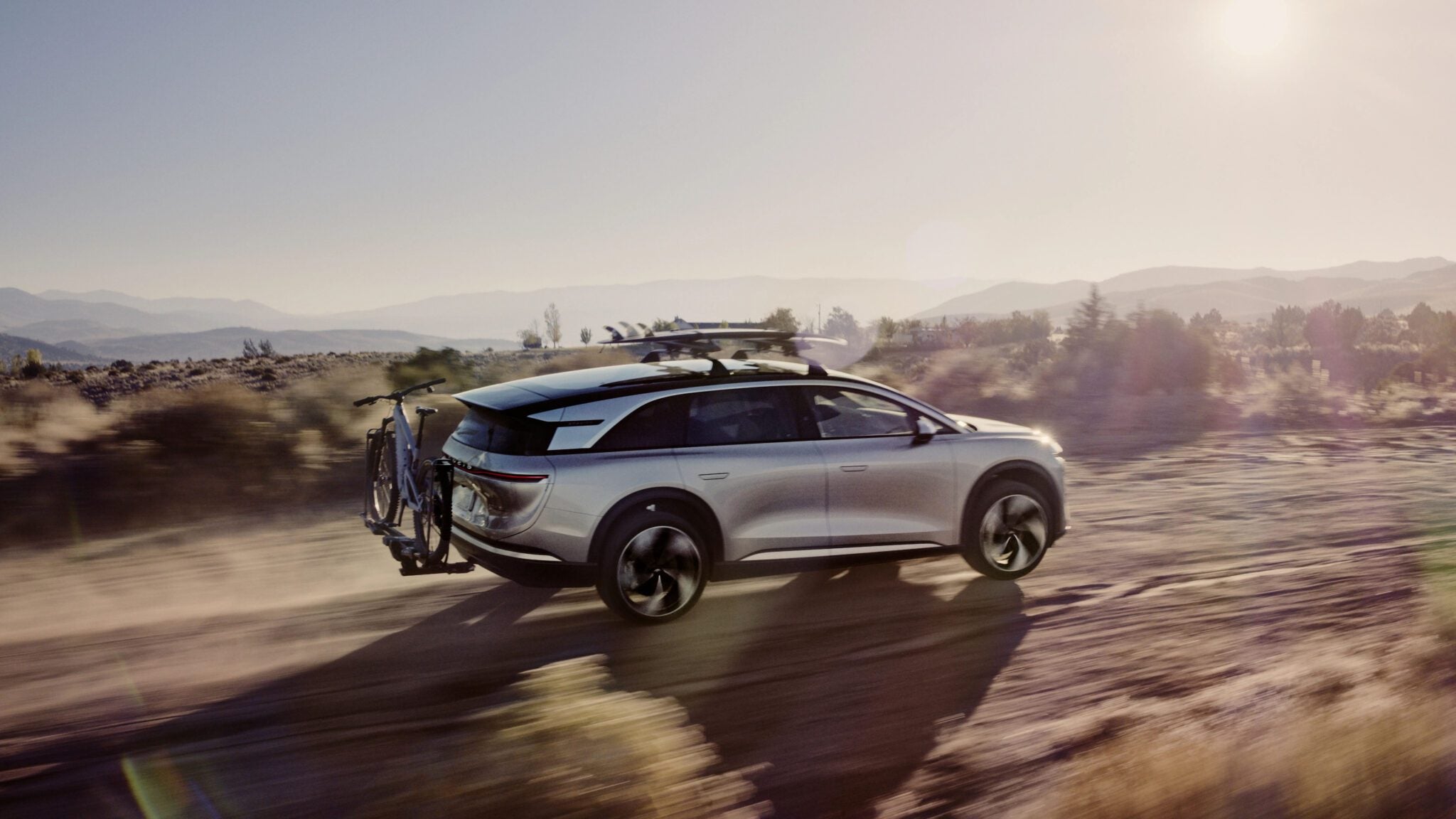 A silver SUV with a roof rack and a bicycle mounted on the rear drives on a dirt road in a desert landscape during sunset.