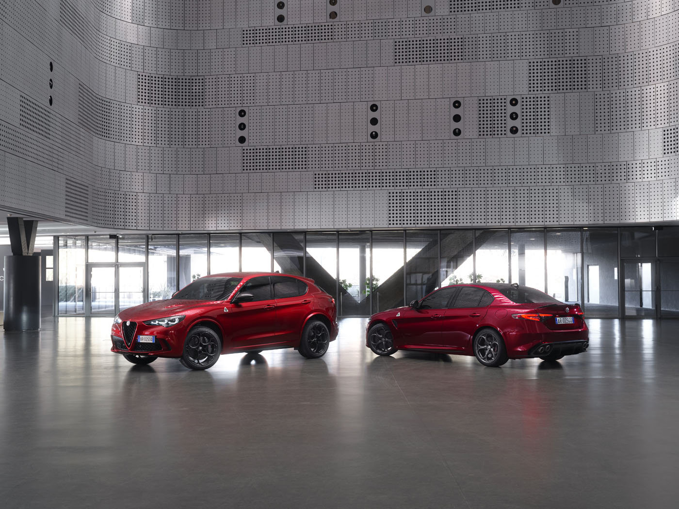 Two red Alfa Romeo vehicles, an SUV and a sedan, are parked inside a modern building with metallic perforated walls and large windows.