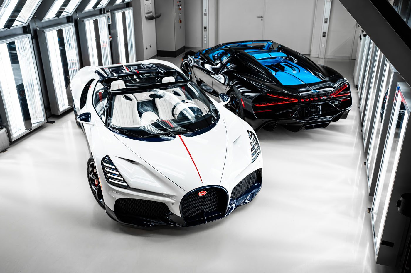 Under bright lighting, two luxury sports cars—a white and a black model—are expertly showcased indoors. These Bugatti masterpieces, including the powerful W16 Mistral, captivate with their sleek, aerodynamic designs and prominent front grilles.