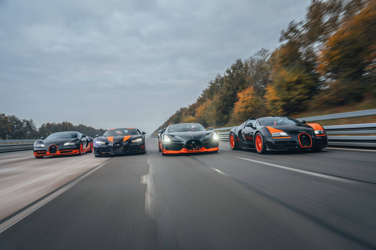 Four Bugatti sports cars, including the renowned W16 Mistral, drive side by side on a highway. Trees line the road under an overcast sky as these high-performance machines hint at their speed record potential.