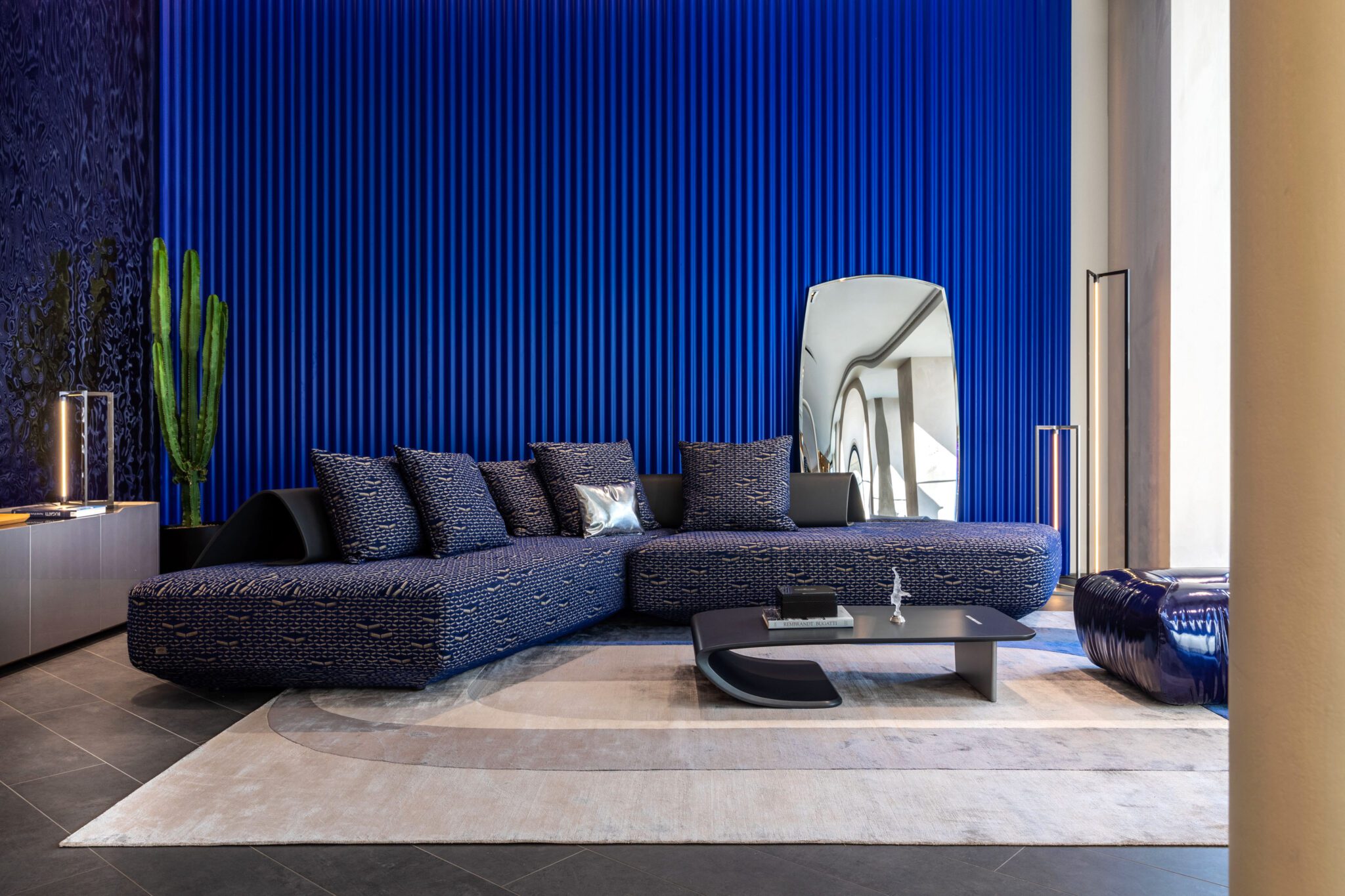 Modern living room with a blue sectional sofa, textured blue wall, cactus plant, abstract mirror, and minimalist decor on a neutral rug from the Paris Home Collection.