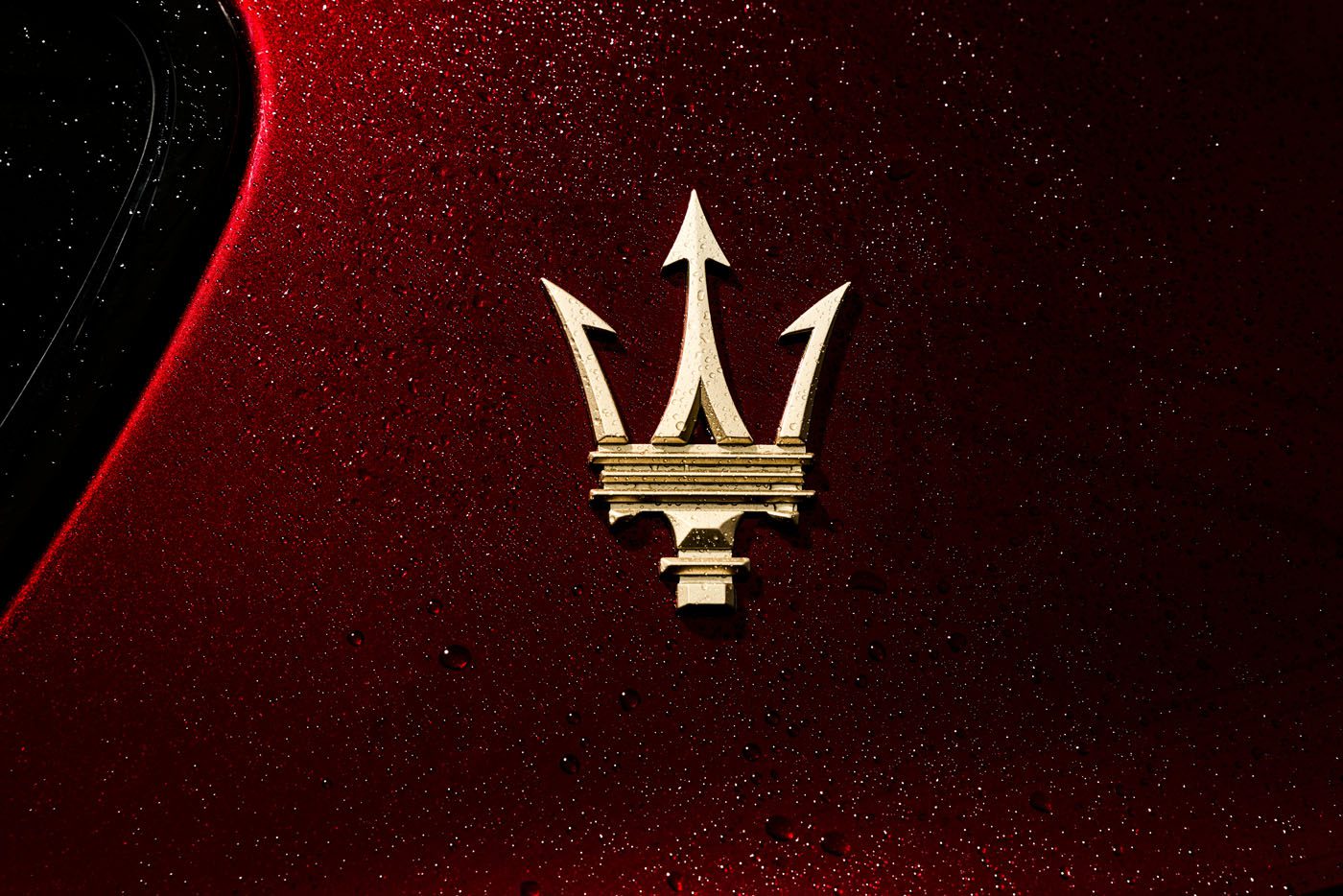 Gold Maserati trident logo on a red, water-speckled car surface.