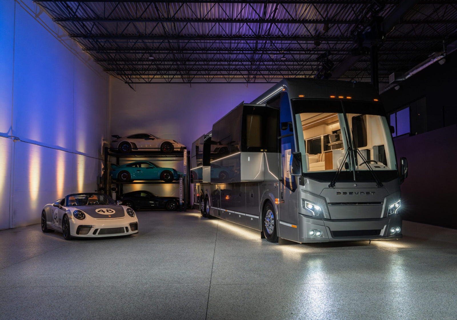 A luxury Off-Grid Prevost Motorcoach by LOKI Coach is parked indoors beside a white Porsche sports car, with additional sports cars stored on racks in the background.