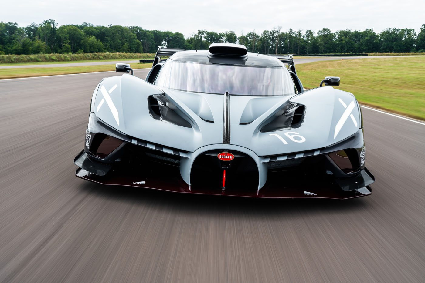 A grey Bugatti Bolide supercar drives on a racetrack, viewed from the front with trees and grass in the background.