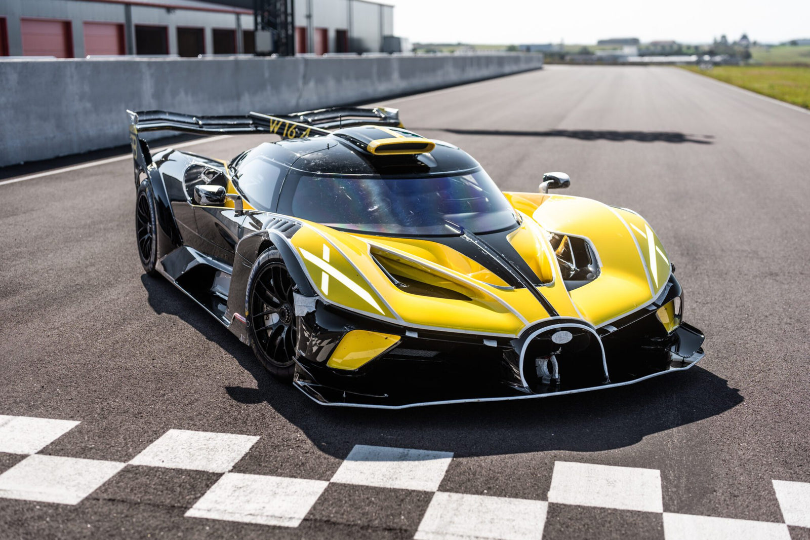 A sleek yellow and black Bugatti Bolide hypercar is parked on a racetrack near distinctive checkered markings, ready for its shakedown test.