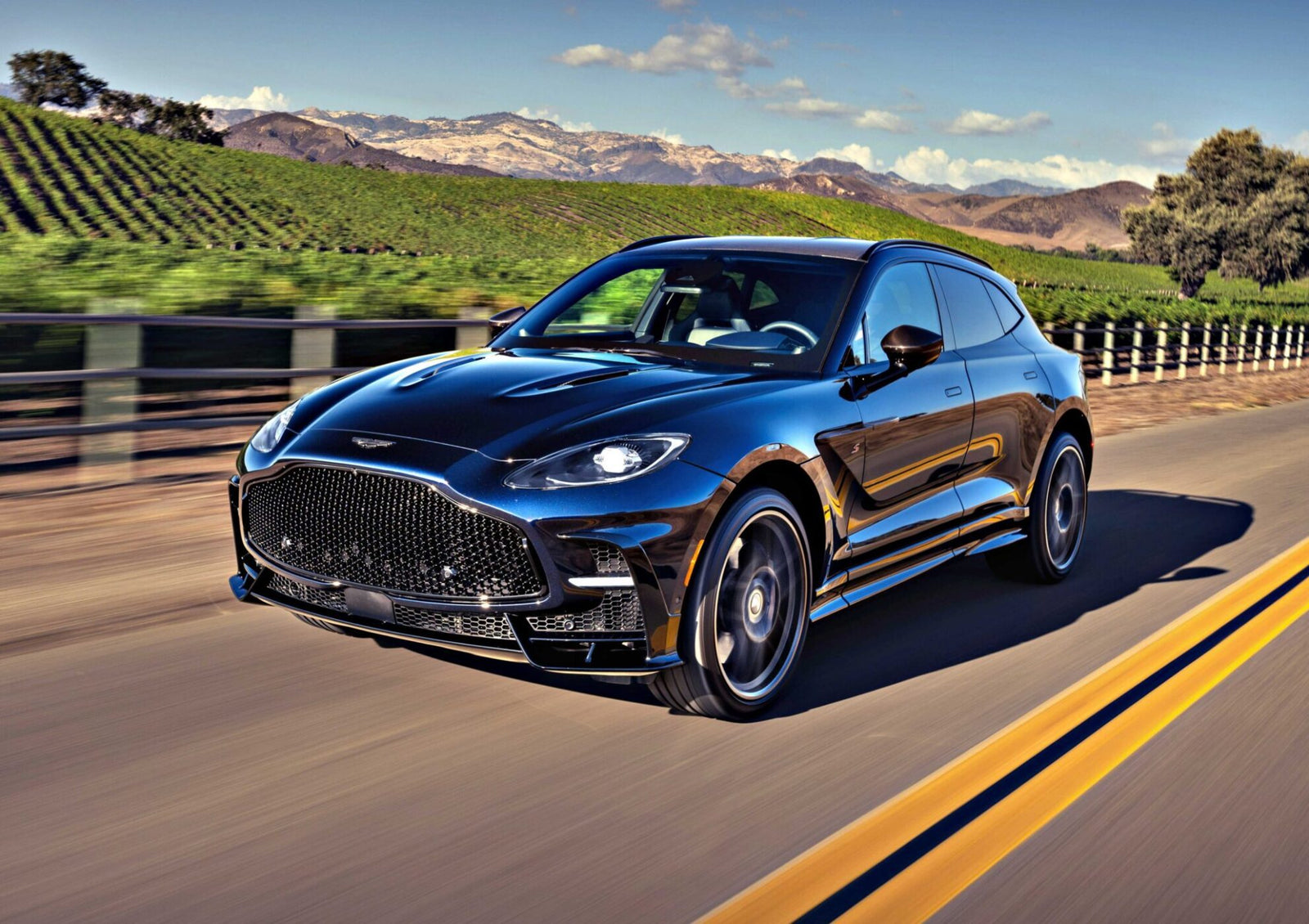 A black 2026 Aston Martin DBX S First Drive: The Best Super SUV Gets Even Better cruises on a paved road past a vineyard, framed by mountains and blue sky.