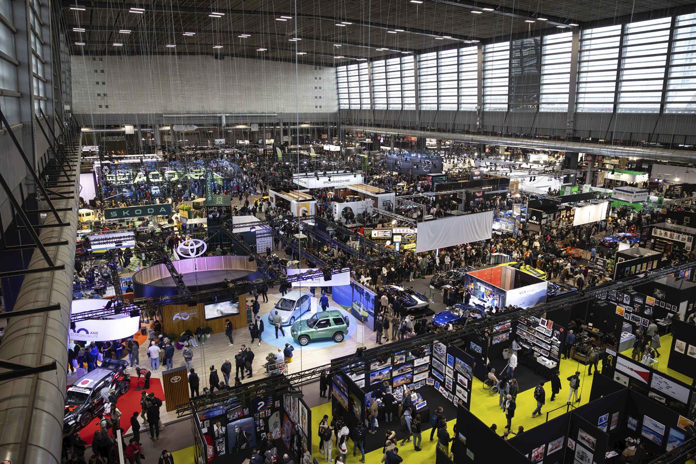 Large indoor car exhibition with numerous booths, display vehicles, and crowds of attendees, reminiscent of the vibrant atmosphere at Rétromobile or a prestigious Auction House like Gooding Christie's, in a spacious, well-lit convention hall.