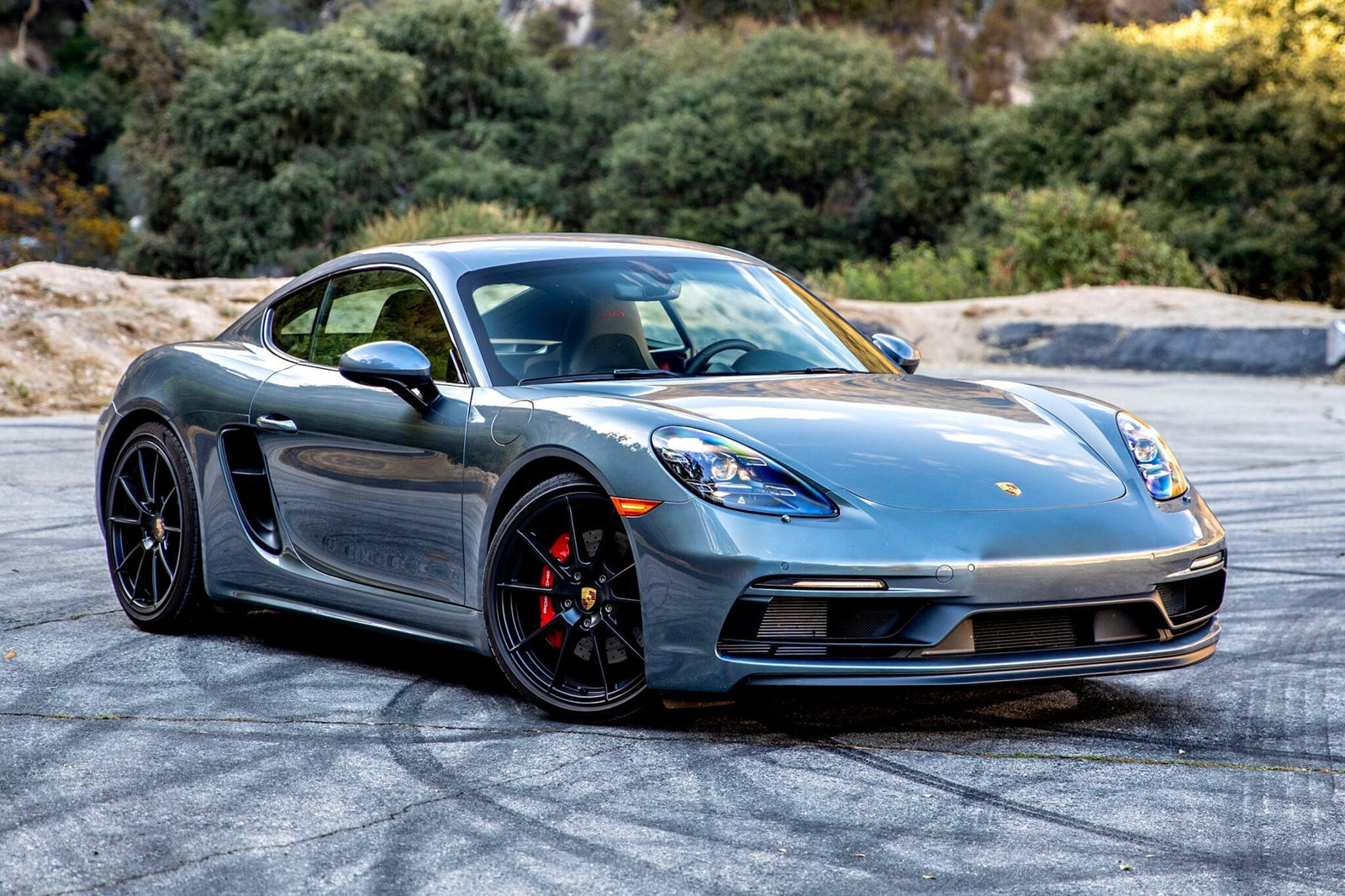 A gray Porsche sports car is parked on a paved area with trees and rocks in the background, echoing the sleek style highlighted in the Review: 2025 Porsche 718 Cayman GTS 4.0 Is Close To Flawless.