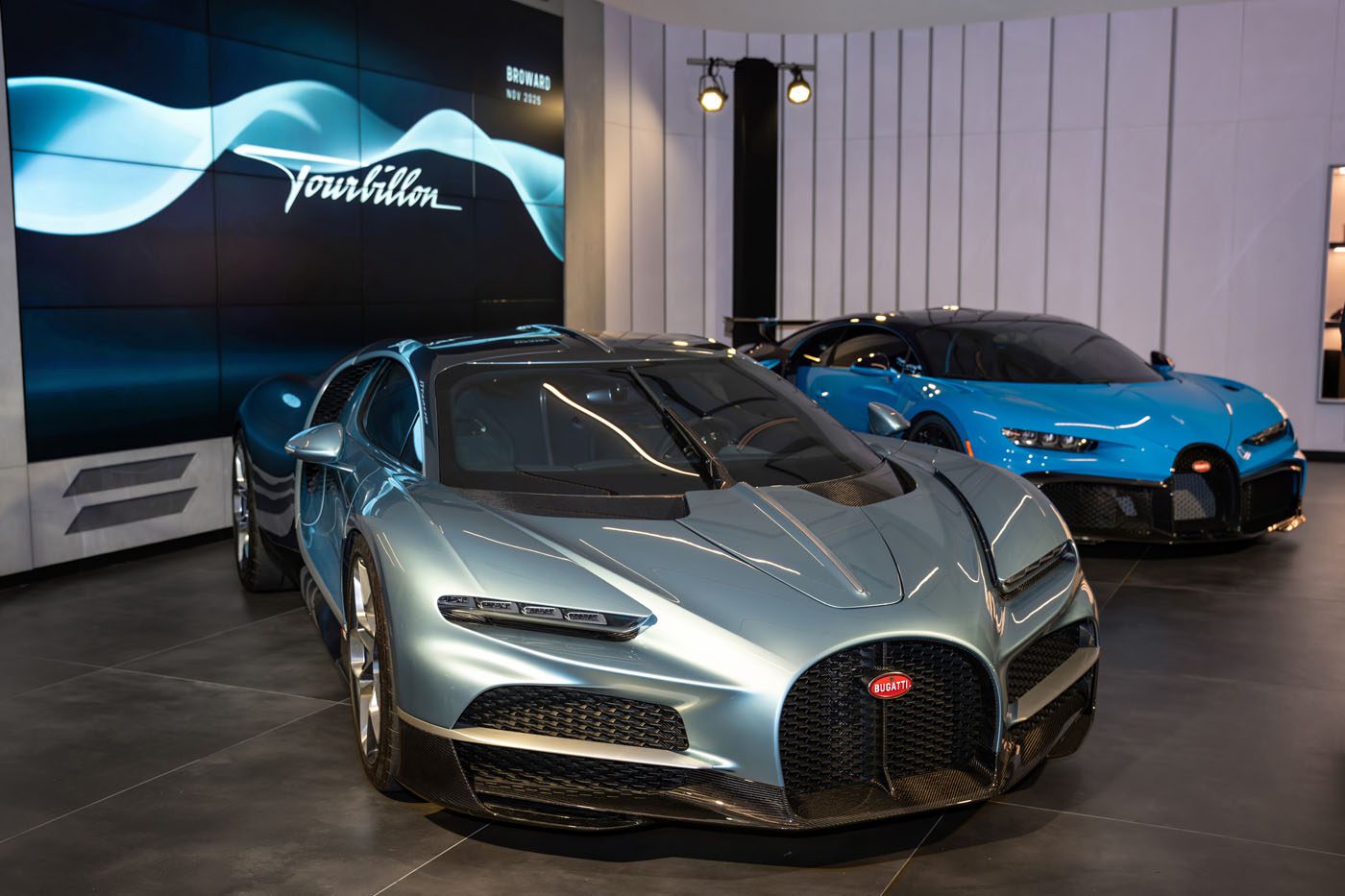 Two Bugatti hypercars, one silver and one blue, are displayed in a modern Bugatti Broward showroom with a digital screen reading "Tourbillon" in the background.