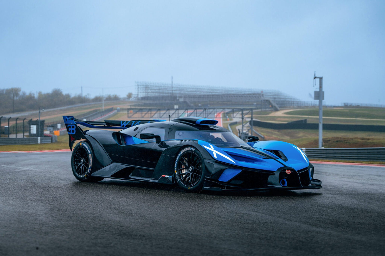 A sleek black and blue Bugatti Bolide is parked on a wet race track under overcast skies, epitomizing the essence of a track hypercar at COTA.
