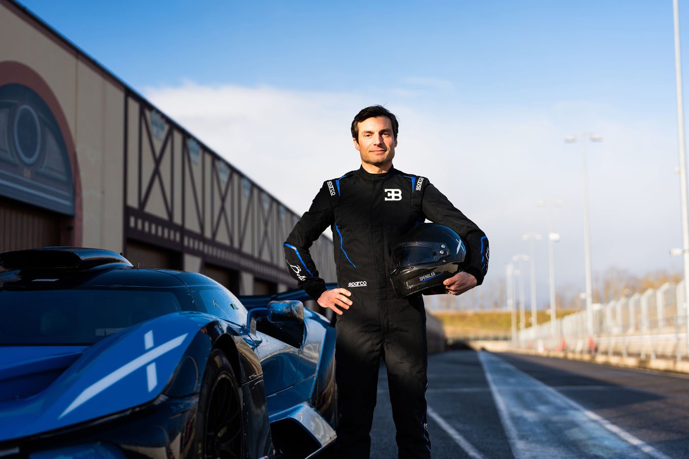 A person in a racing suit, possibly a Pilote Officiel, stands beside a blue Bugatti on the racetrack, holding a helmet. The backdrop reveals a sunny day with the building facade gleaming in the distance.