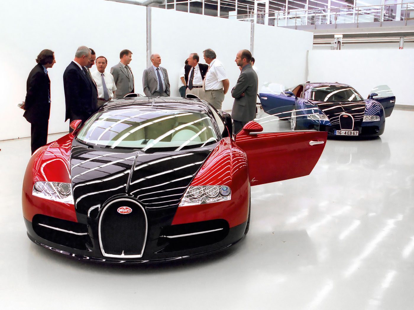 A group of men in business attire stand near two Bugatti Veyron cars, famed for their W16 engines, in a clean, modern showroom with white walls and glossy floors.