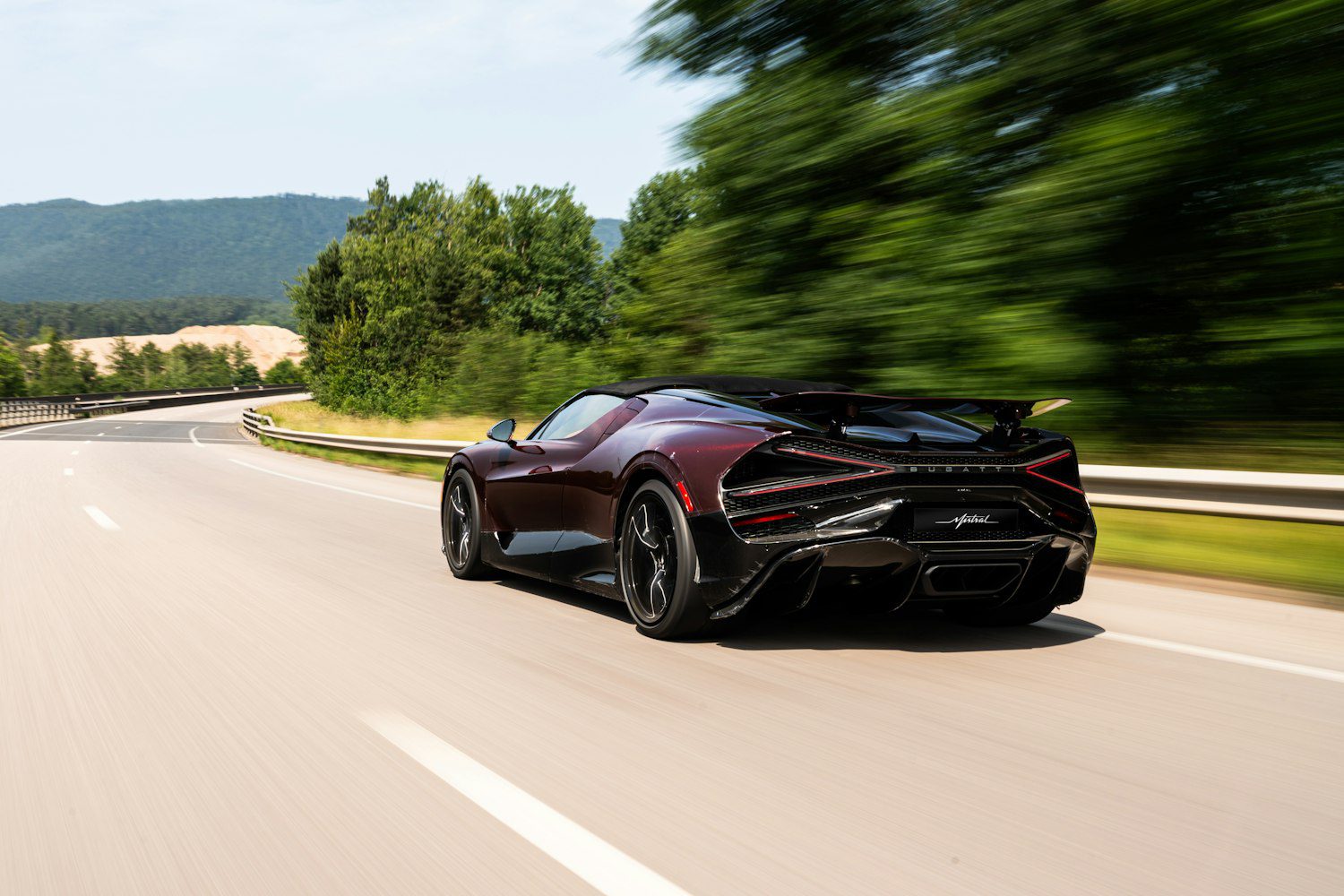 A sleek black Bugatti Mistral speeds down an empty highway bordered by green trees, the blurred background capturing its motion.