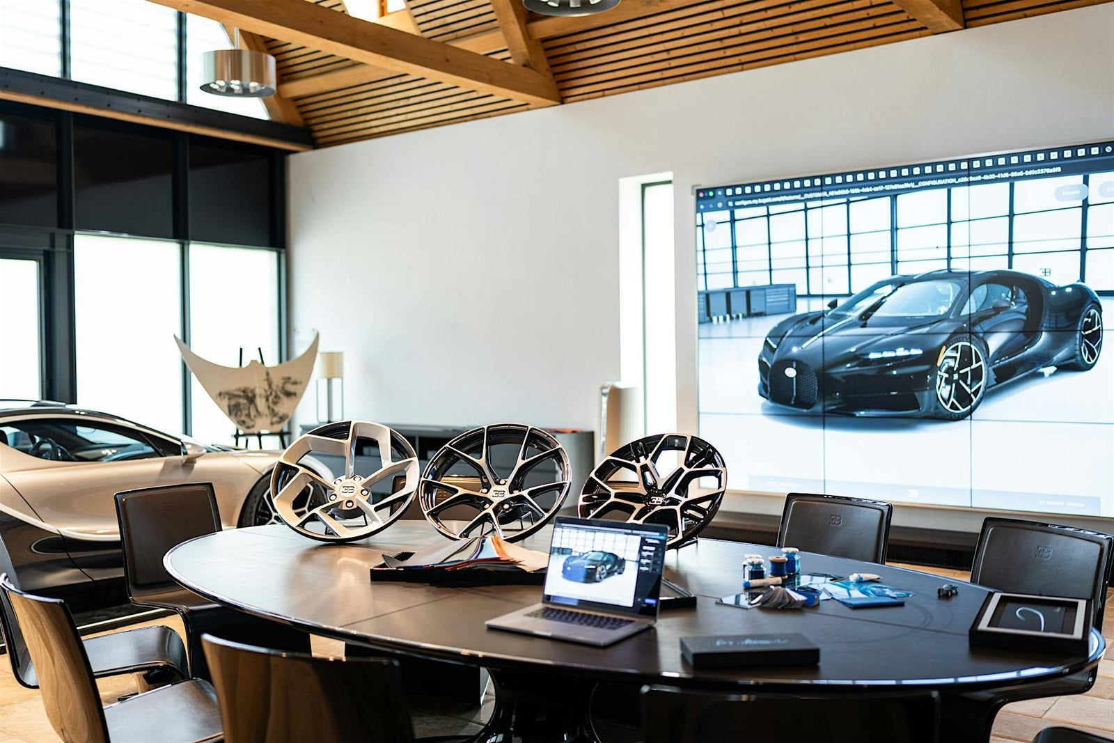 A conference room with car rims on a table, a laptop, and a large screen displaying a Bugatti Tourbillon in the background—perfect for configuring your next luxury experience.