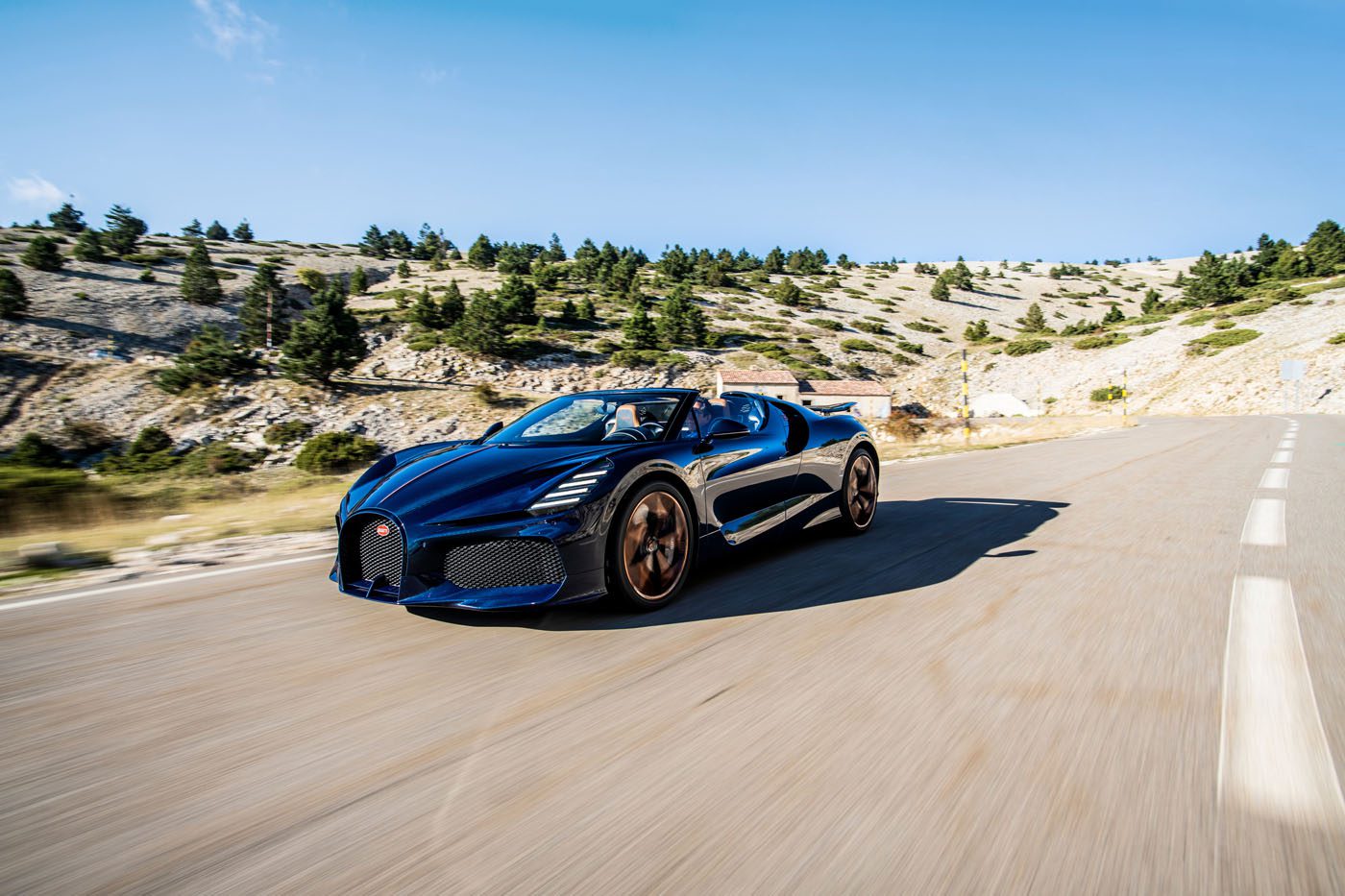 A blue Bugatti hypercar with tan wheels speeds along a scenic road, hills and trees lining the background under a clear blue sky.