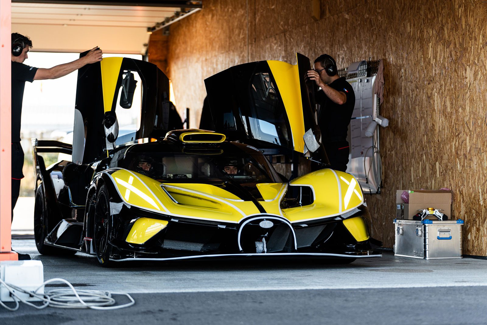 A yellow and black Bugatti Bolide with its butterfly doors open is being prepared by two people in a Miller Motorcars garage.