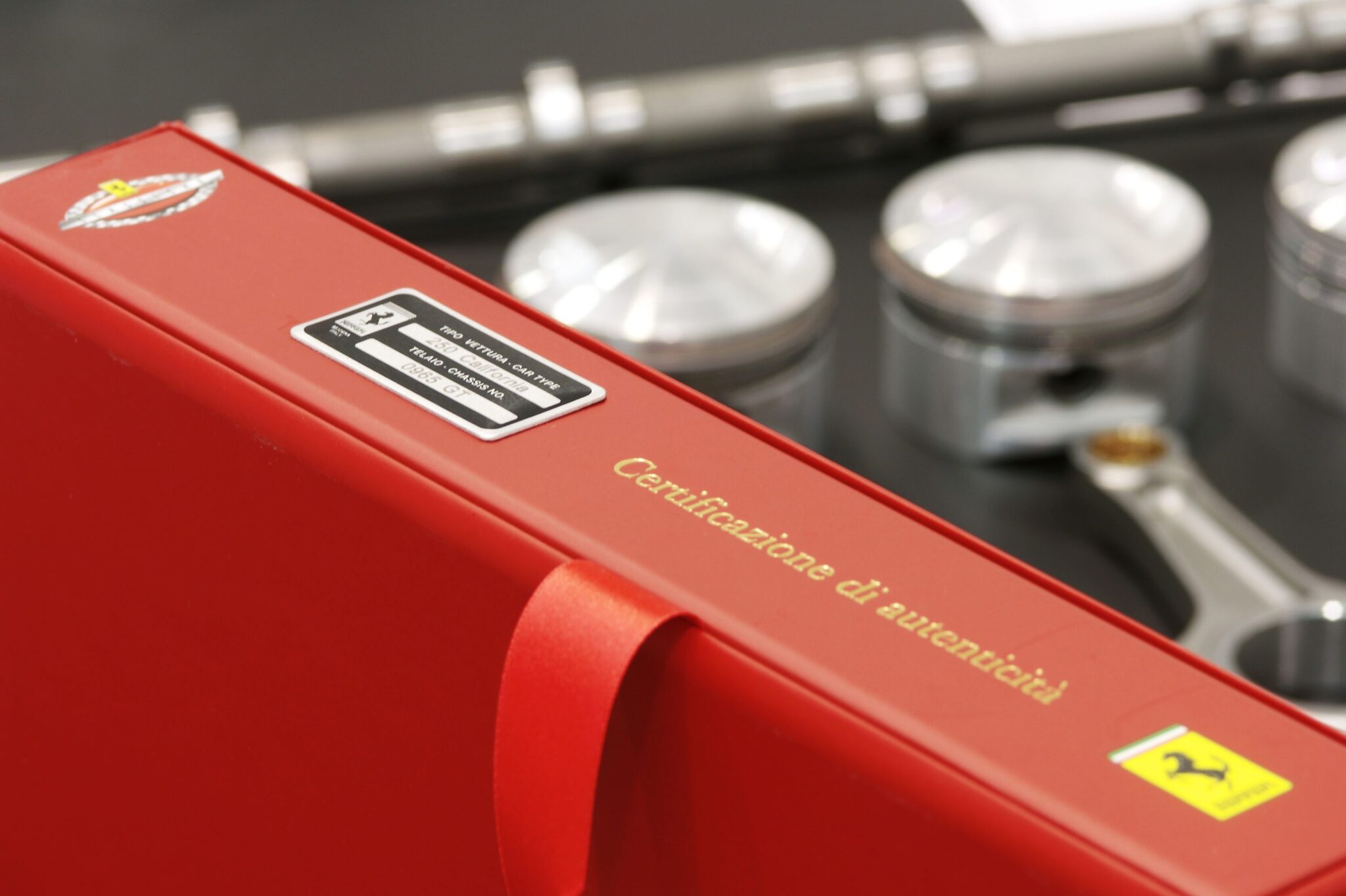 A red binder labeled "Certificazione di autenticità" with Ferrari logos is shown in front of automotive engine parts, including three pistons and a wrench.