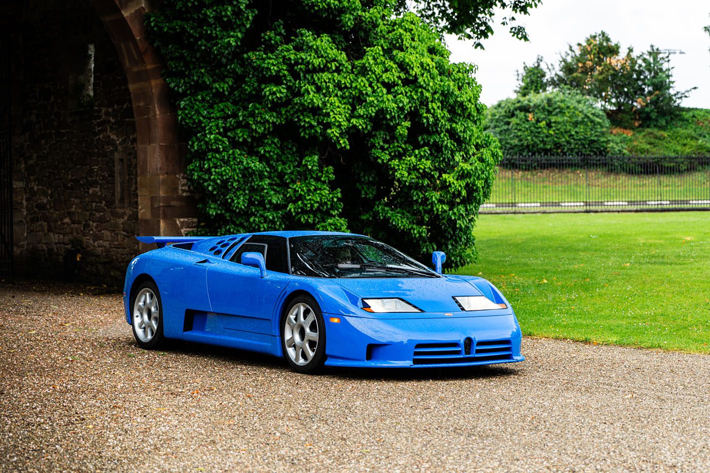 A blue Bugatti La Maison Pur Sang sports car is parked on a gravel path near a stone wall, with grass and trees in the background.