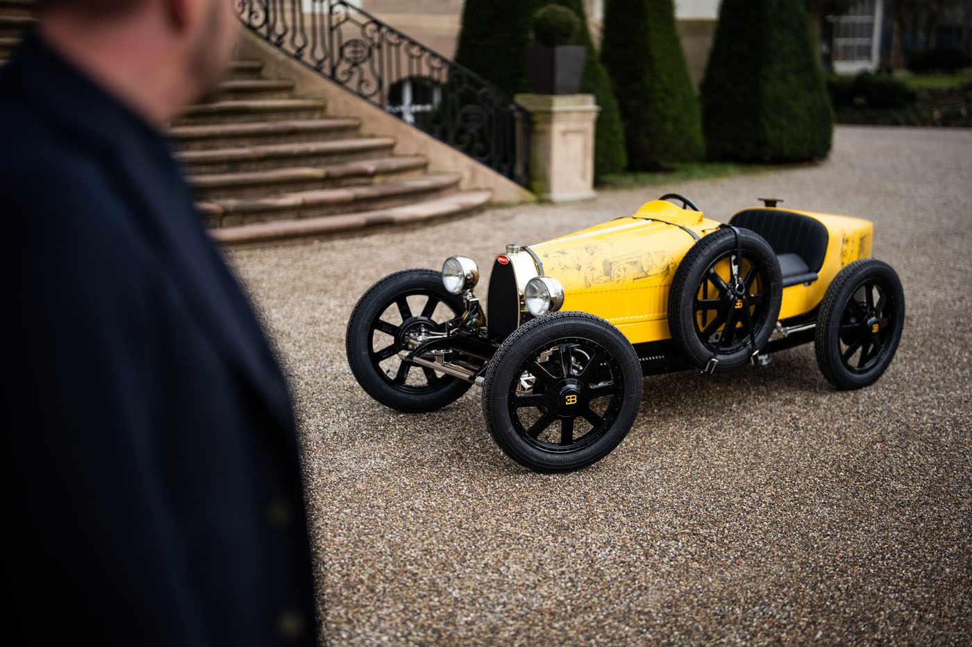 A small, vintage-style yellow car resembling a Bugatti Baby II with black wheels is parked on a gravel driveway. A person stands in the foreground, turned slightly toward the car.