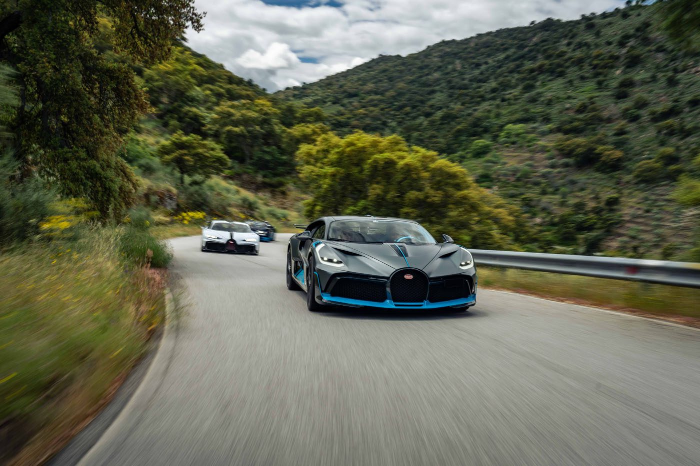 Three sports cars drive on a winding mountain road surrounded by greenery and hills under a partly cloudy sky.