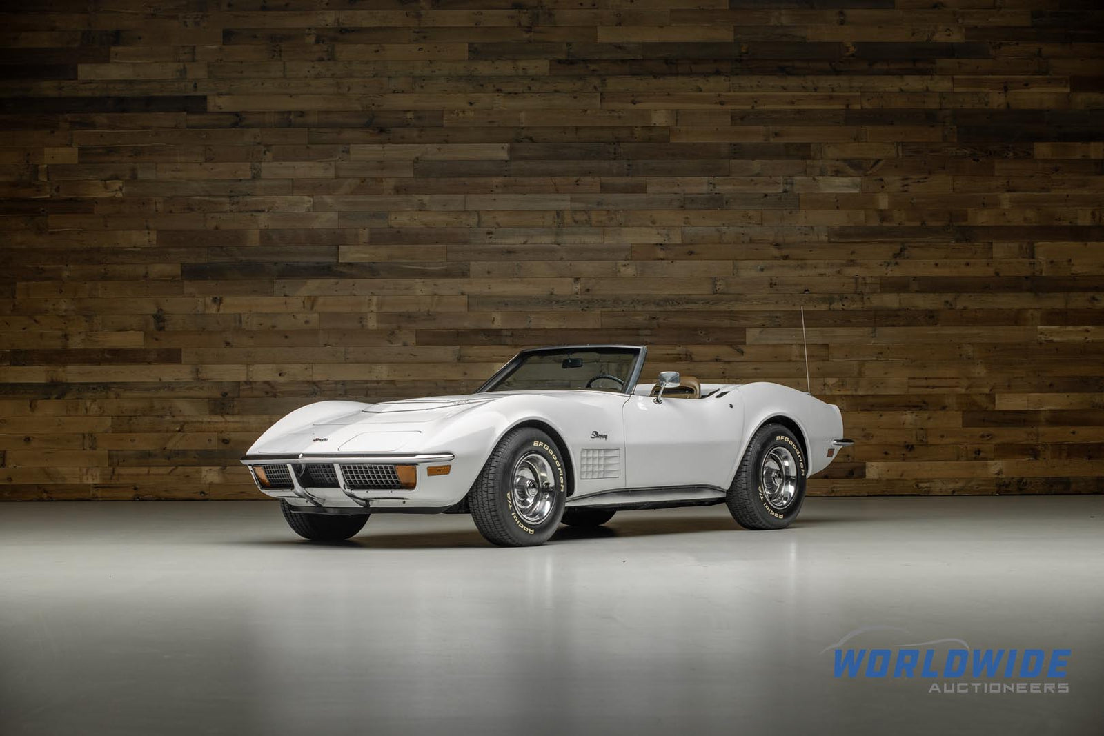 A white vintage convertible with the top down is parked indoors on a smooth floor, set against a wooden wall. The "Worldwide Auctioneers" logo hints at an Enthusiast Auction featuring a diverse lineup of classic cars.