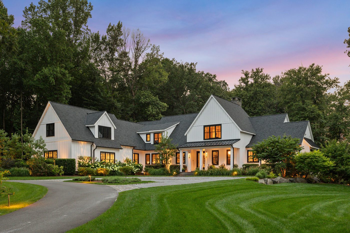 A large, modern farmhouse-style home with white siding and dark trims is surrounded by trees and a manicured lawn at dusk. A curved driveway leads to the entrance.