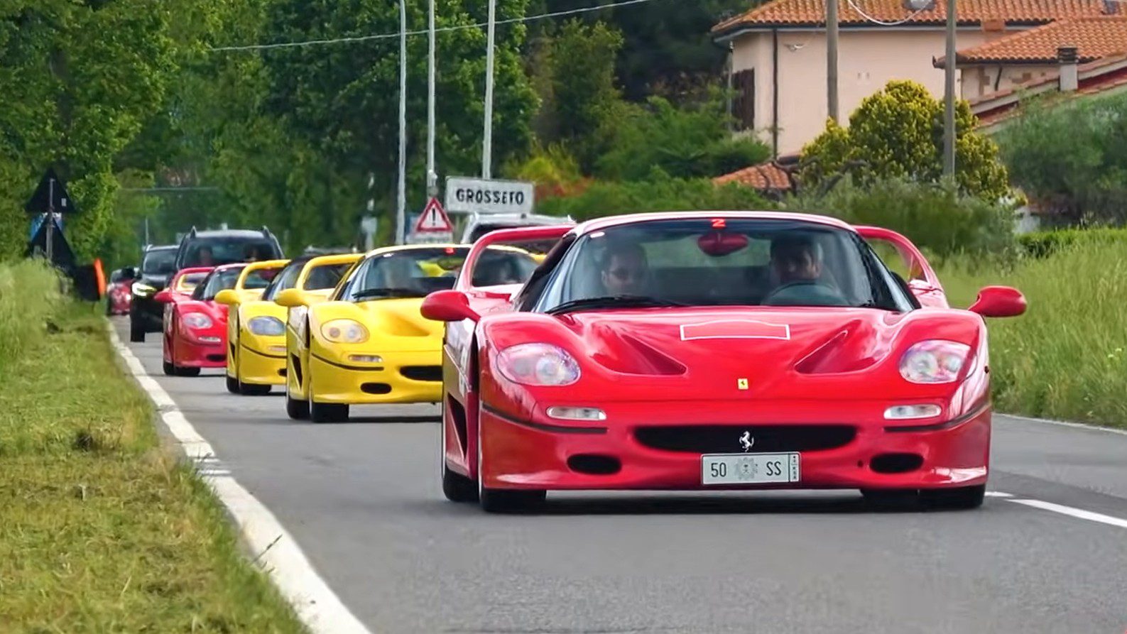 A line of red and yellow Ferrari F50 sports cars drives down a rural road bordered by grassy fields and houses, celebrating the Legacy Tour’s 30th Anniversary.