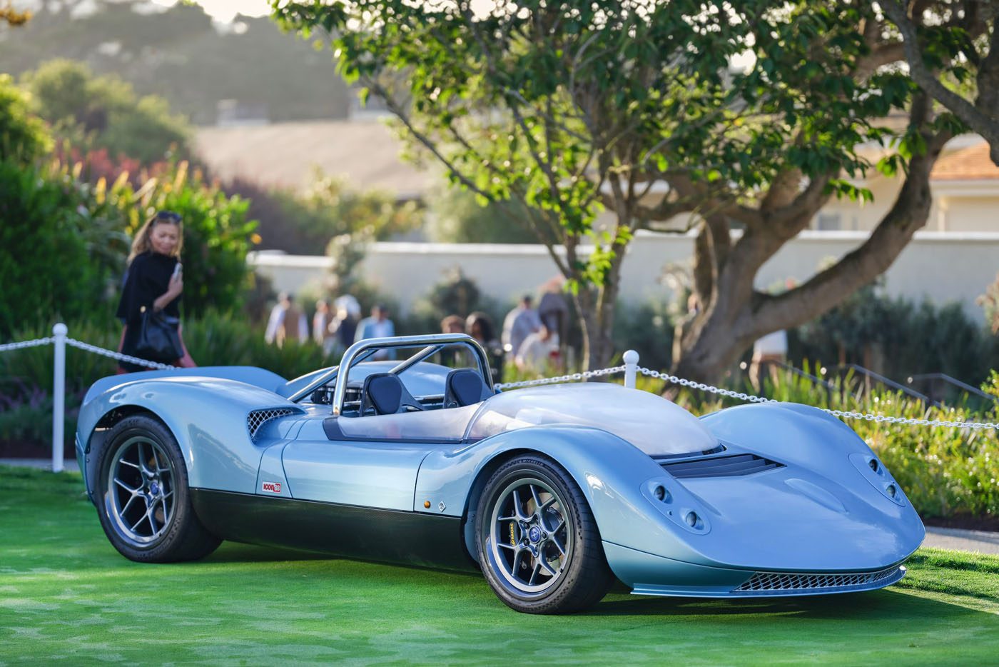 A sleek, light blue vintage-styled sports car is displayed on a green lawn with trees and people blurred in the background.