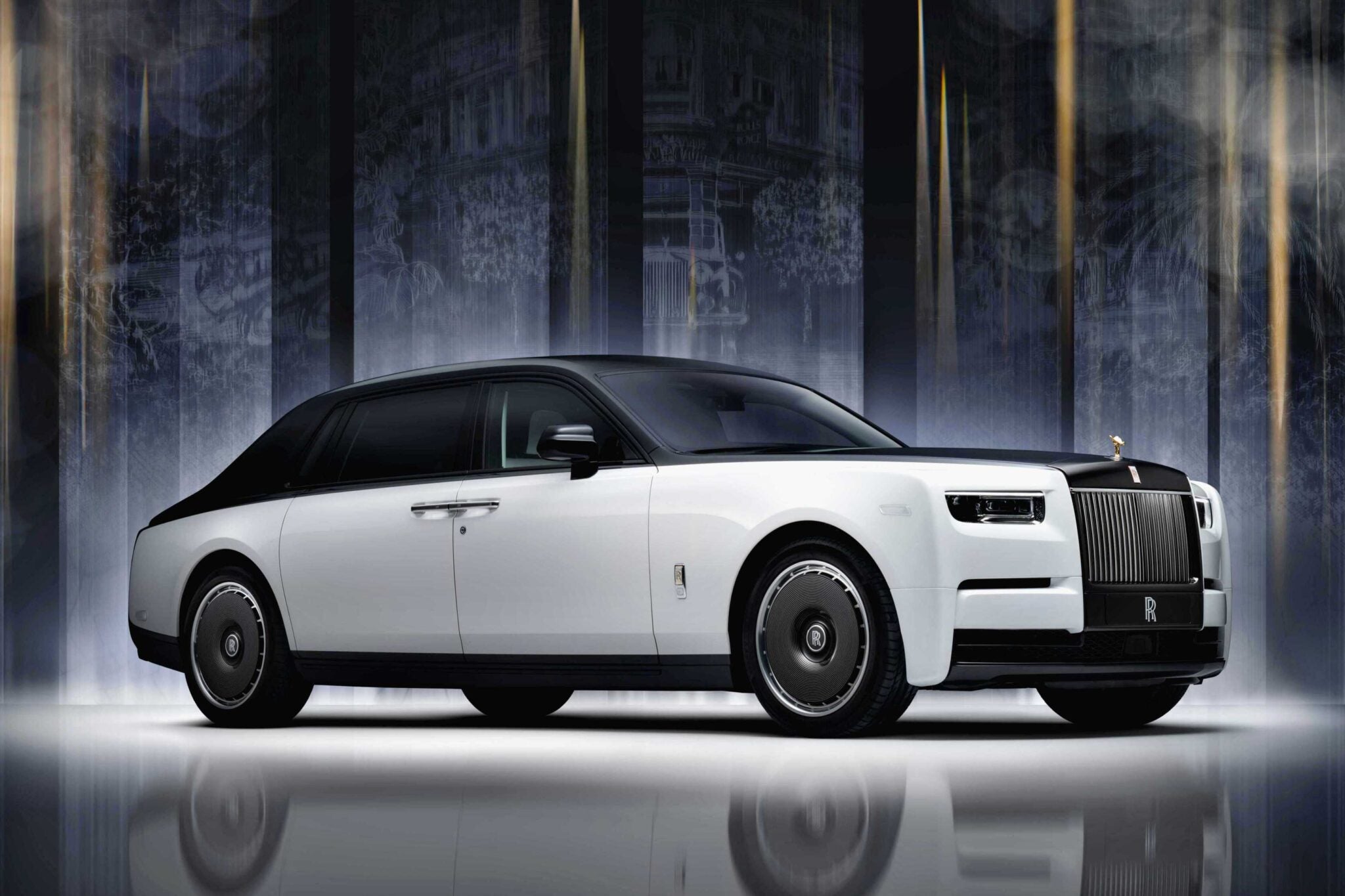 A white Rolls-Royce Phantom luxury sedan with black accents is parked indoors against a dark, illuminated background, celebrating Rolls-Royce Honors 100 Years Of Phantom With Groundbreaking Private Collection.