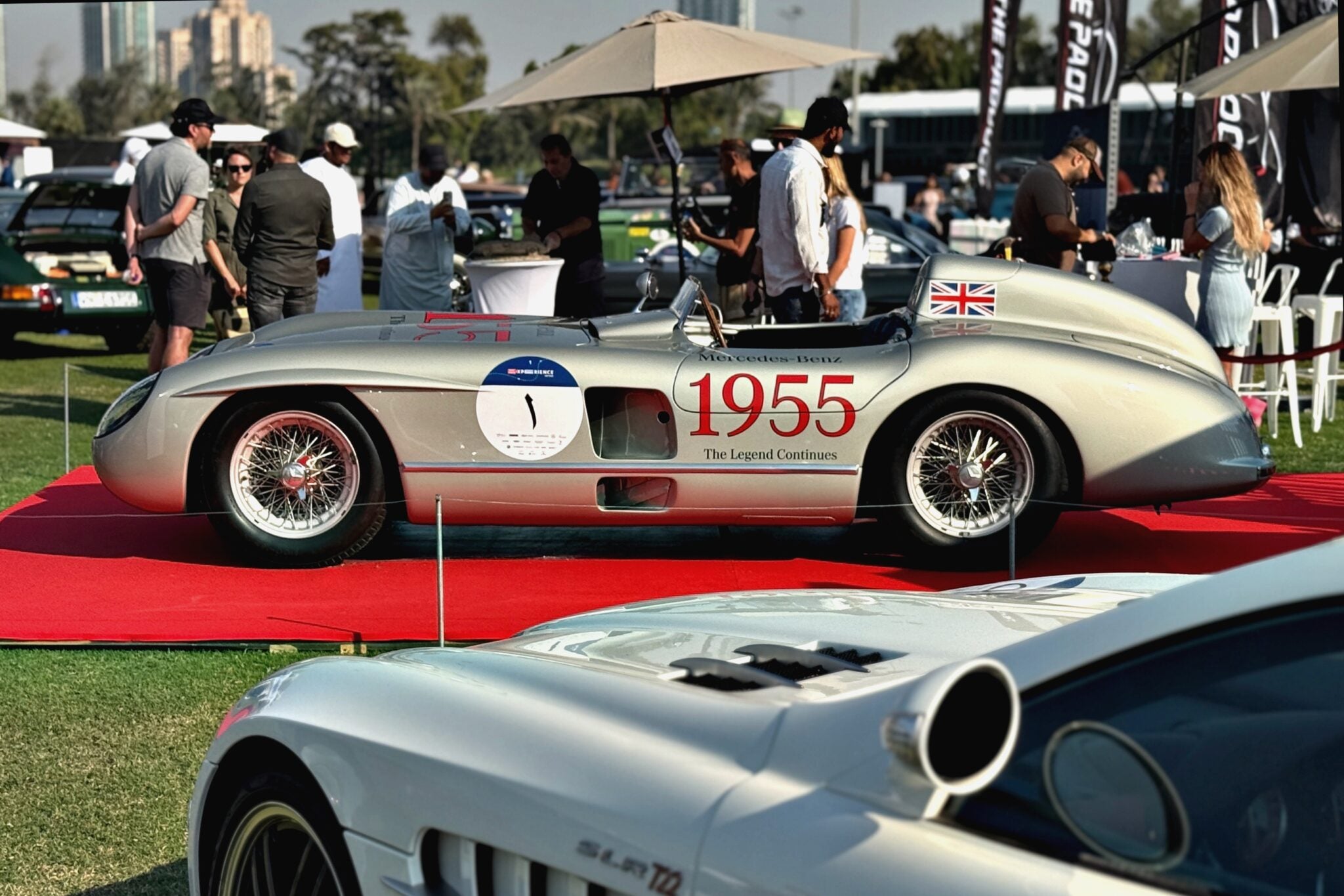 A classic silver race car with "1955" on the side is displayed on a red carpet at an outdoor car show during the 1000 Miglia Experience UAE 2025: Opening Day Highlights, with people and other cars in the background.