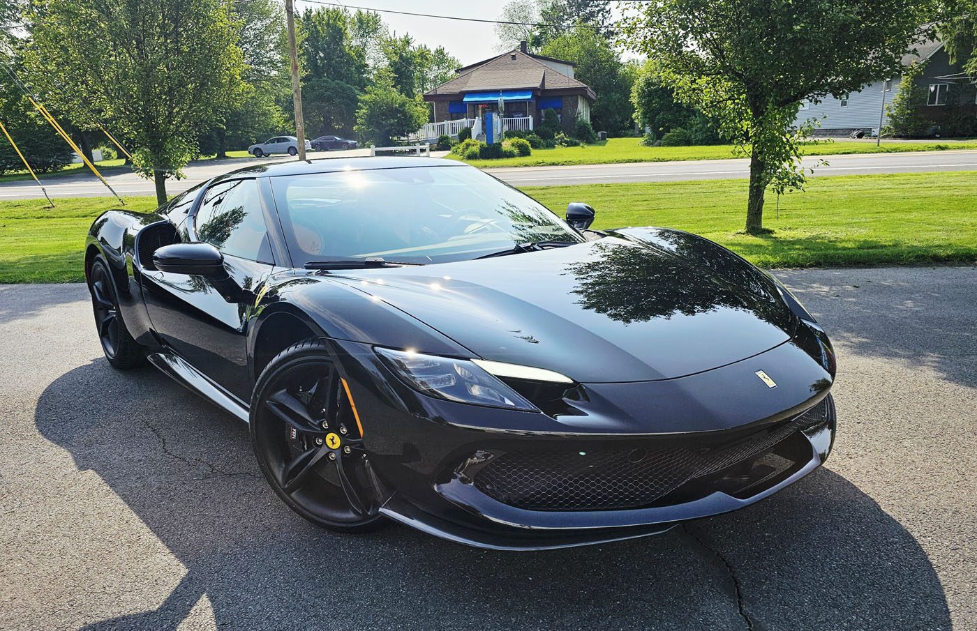 A sleek black Ferrari 296 GTB sports car is parked on a paved driveway, enjoying the Royal Treatment from WrapKings, with trees, grass, and houses in the background on a sunny day.