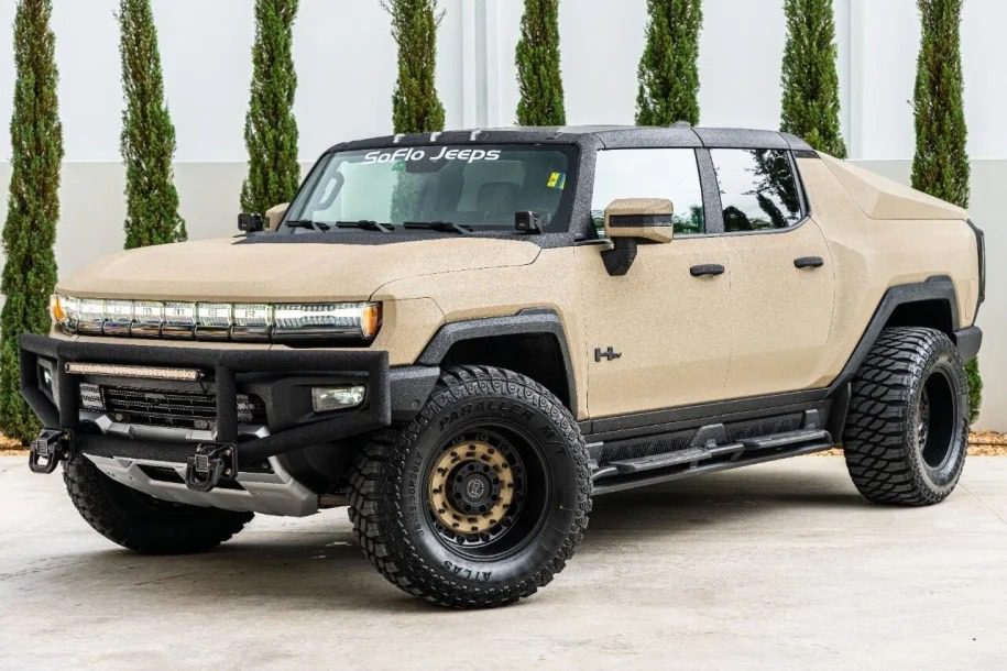A tan customized Hummer EV truck with large off-road tires is parked on pavement in front of tall green shrubs—an impressive example from Unique Creation Garage's Custom Cars For Sale.