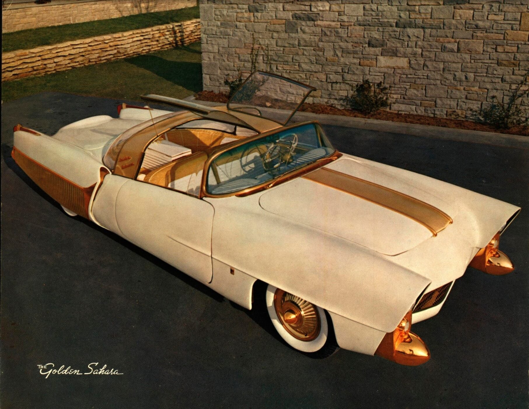The Golden Sahara II, a vintage white and gold convertible with a transparent roof, is parked on a driveway near a stone wall.