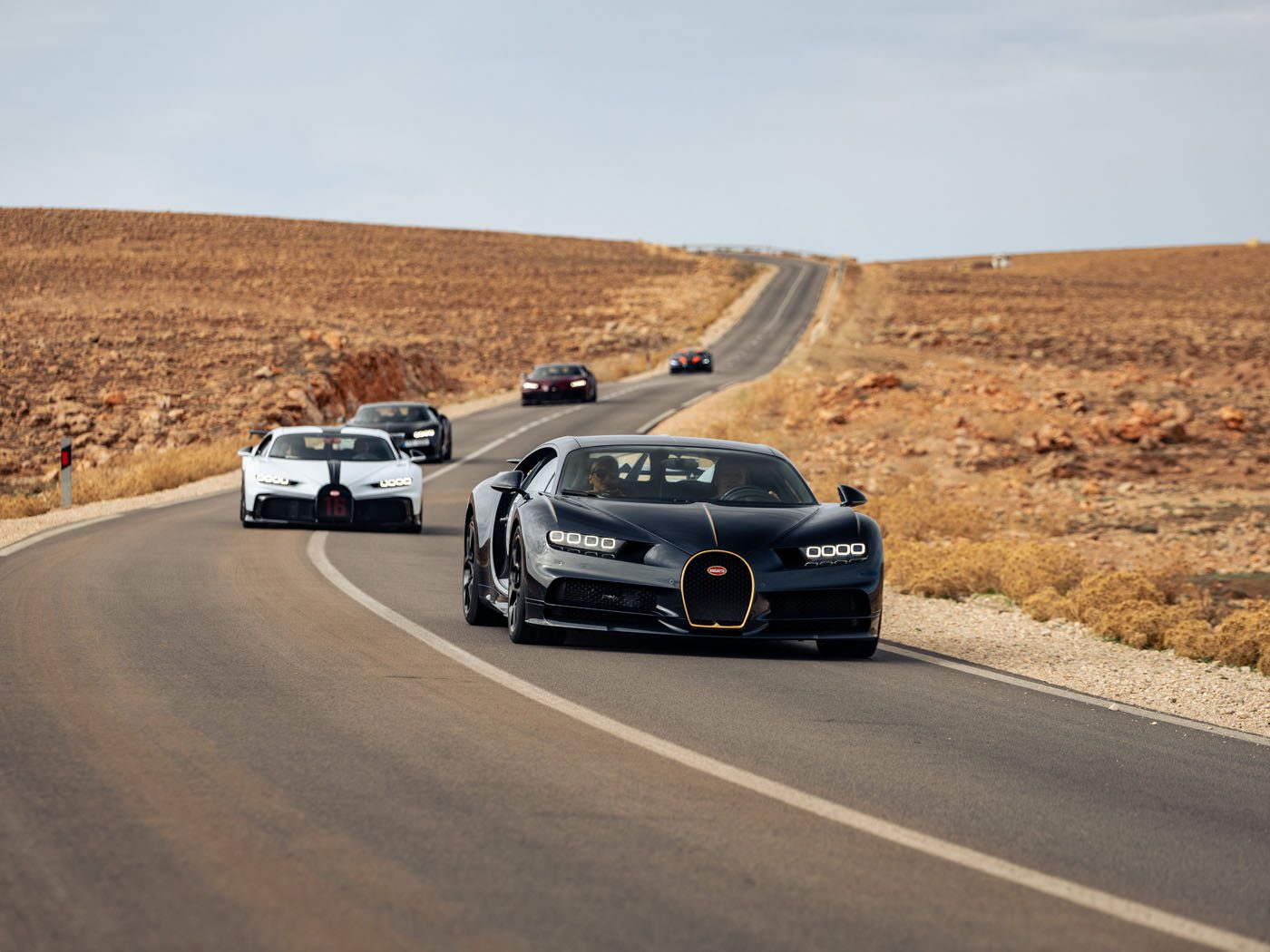 A line of luxury sports cars, including a sleek Bugatti hypercar, cruises along a rural road amidst the stunning desert landscape of Morocco.