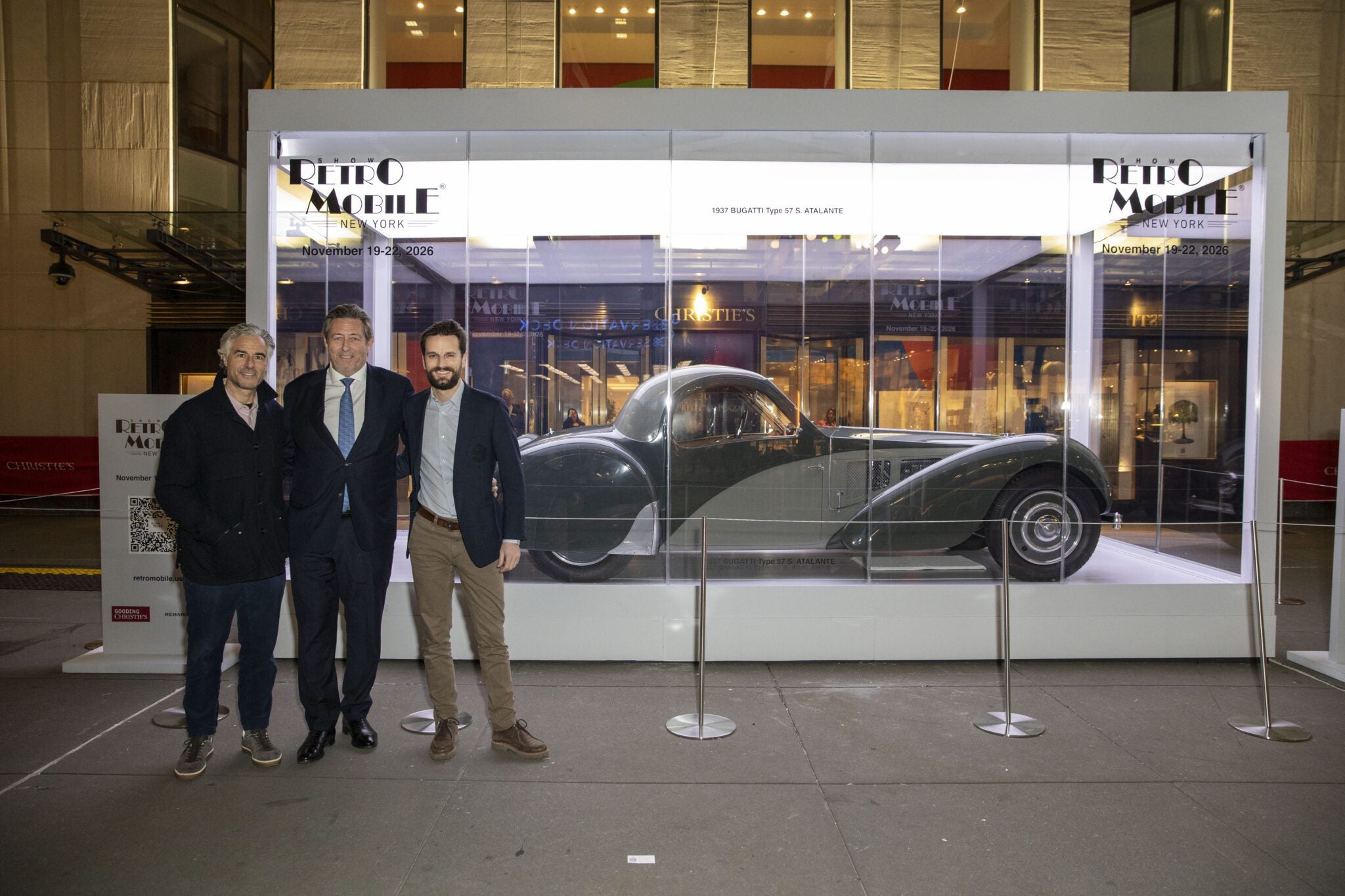 Three men stand before a glass case featuring a 1937 Bugatti Type 57S Atalante at the Rétromobile New York outdoor exhibition, where ticket sales are brisk.