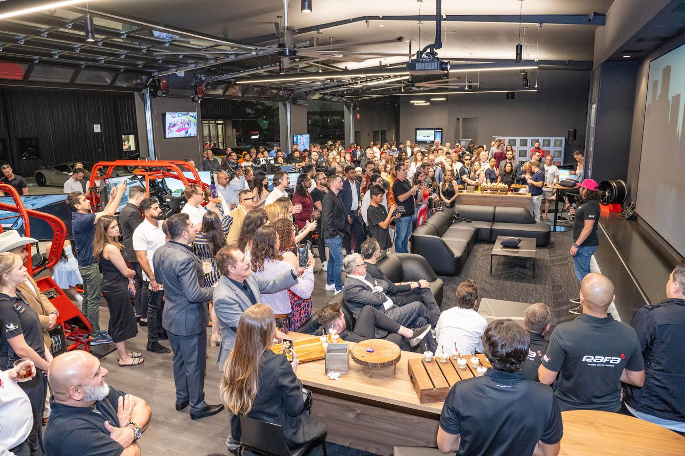 A large group gathers indoors, some seated and others standing, watching a speaker address the crowd in a modern, automotive-themed Houston event space hosted by RAFA Racing Club.