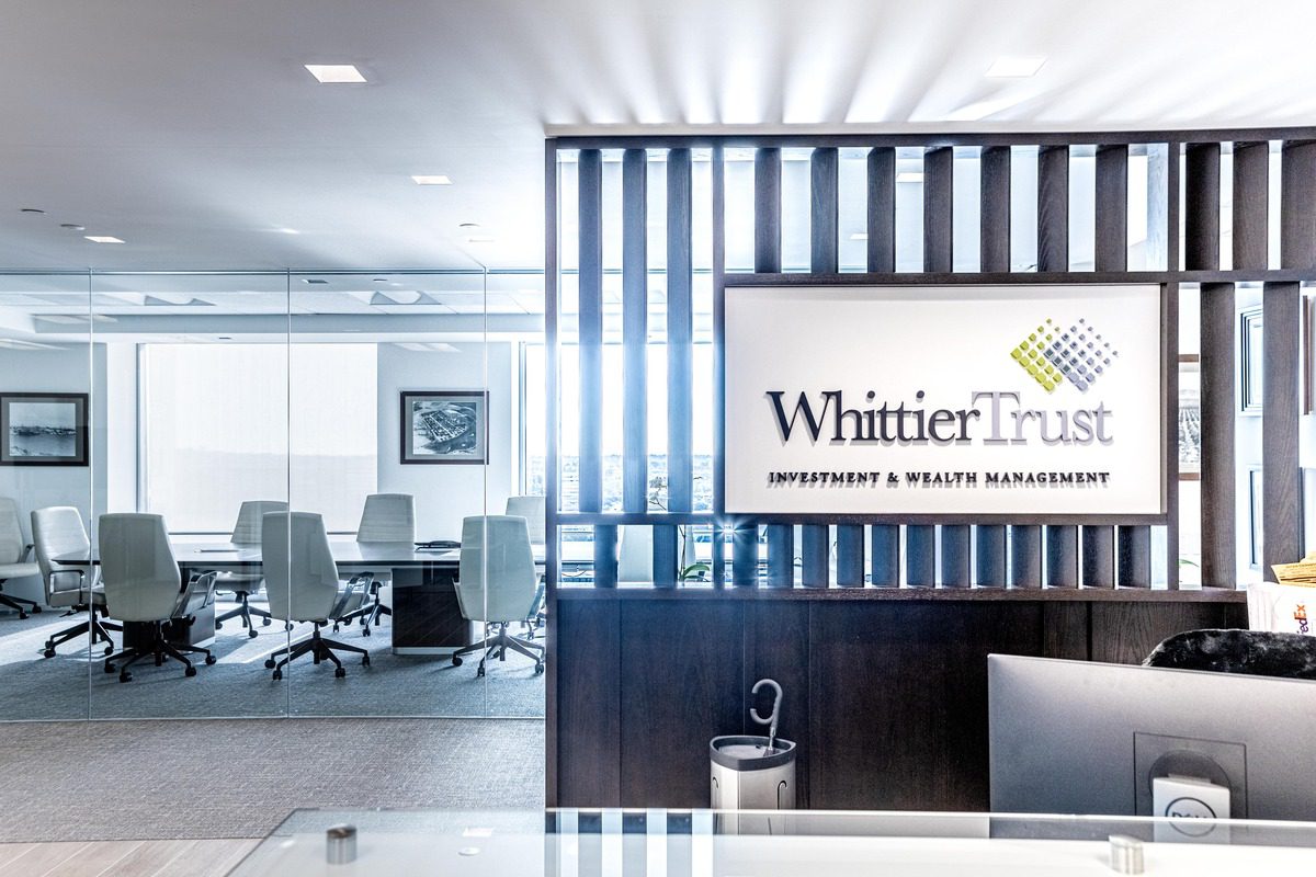 Modern office reception area with a sign reading "Whittier Trust Wealth Management" and a glass-walled conference room with white chairs in the background.