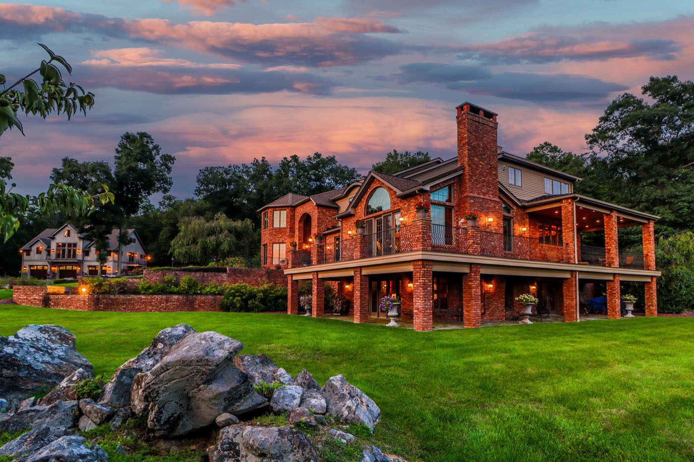 A large two-story brick house with a wraparound patio and outdoor lighting sits on a green lawn at sunset, offering the charm of a mountain-view country estate, with trees and another house in the background.