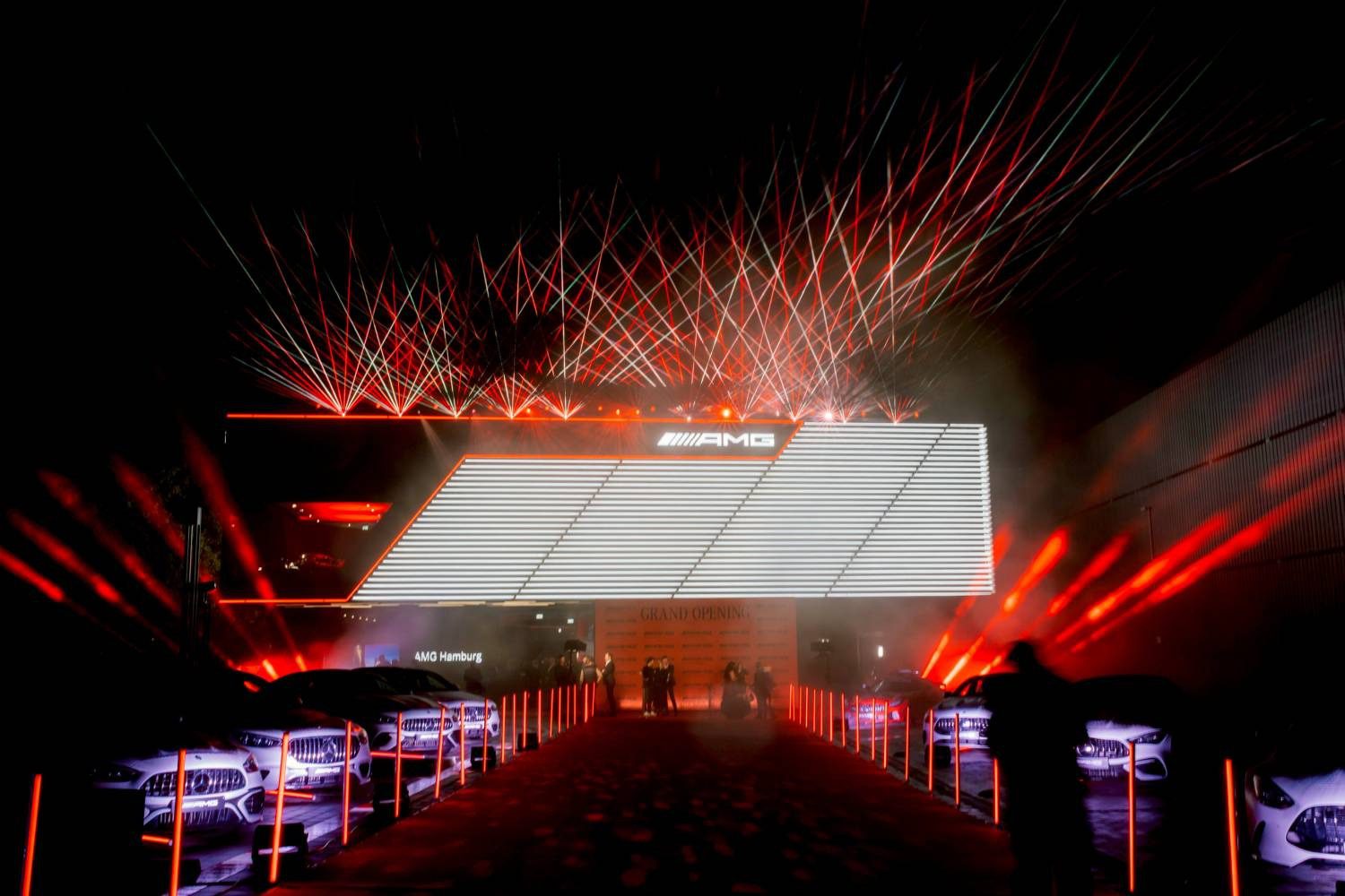 A brightly lit AMG Brand Center event entrance in Hamburg at night with red lasers, illuminated signage, and cars lined up on both sides of the walkway—a true showcase of Germany’s automotive excellence.