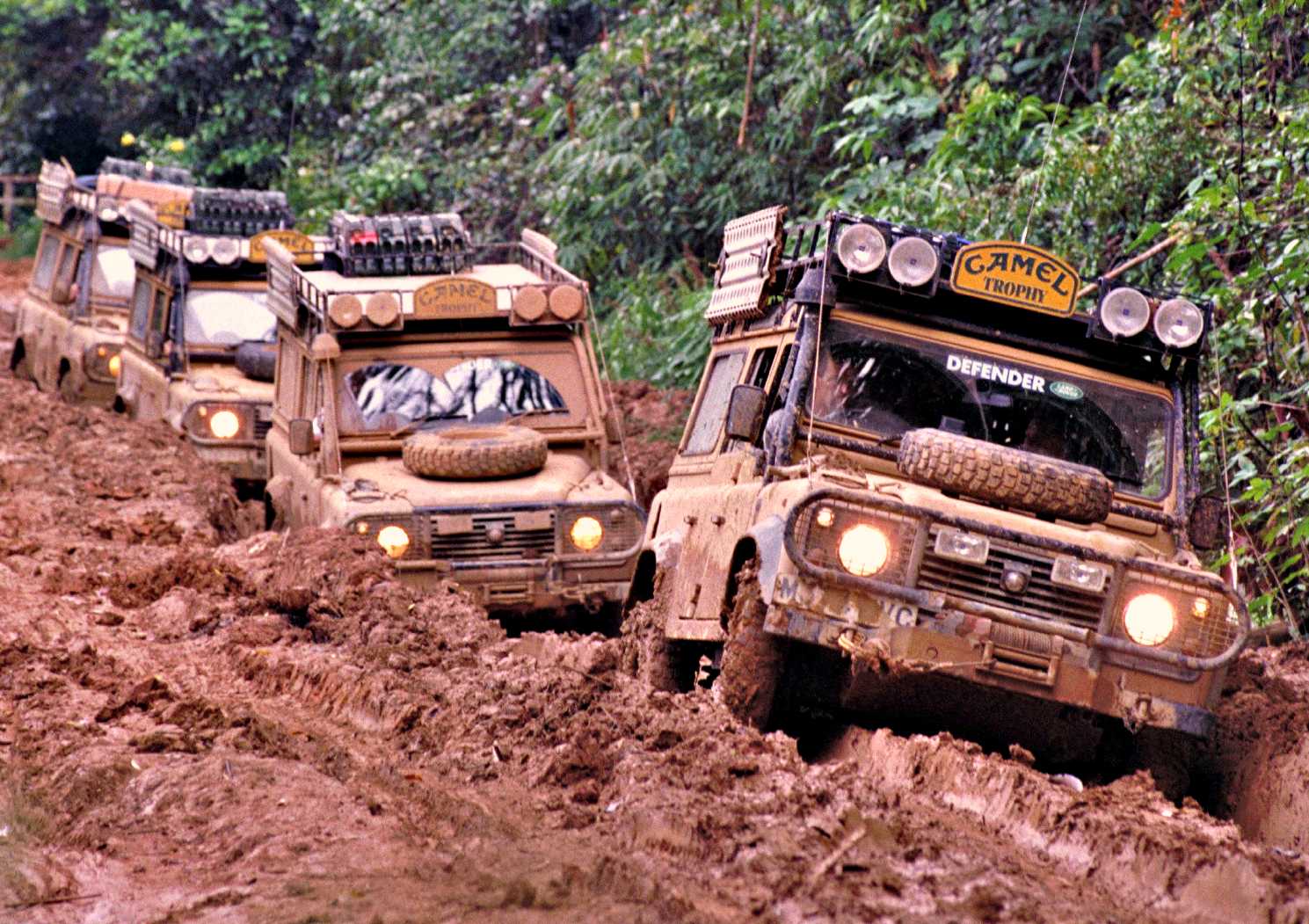 Three mud-covered Land Rover Defenders, true historic expedition icons, drive in a line through a narrow, muddy off-road track surrounded by dense green vegetation.