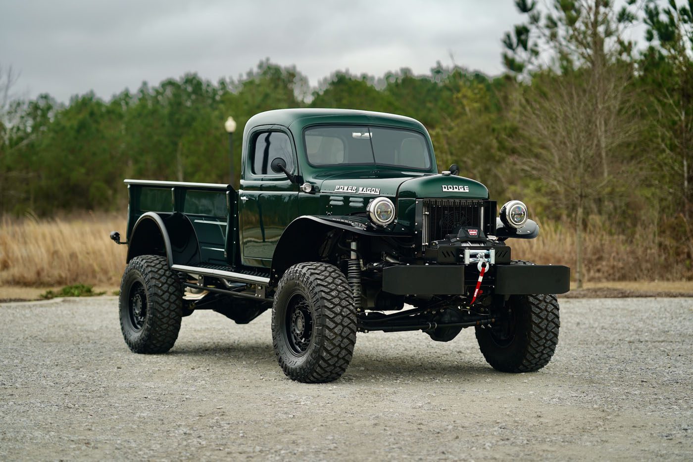 A vintage 1946 Timberline Green Dodge Power Wagon truck is parked on a gravel surface, with trees creating a serene backdrop.