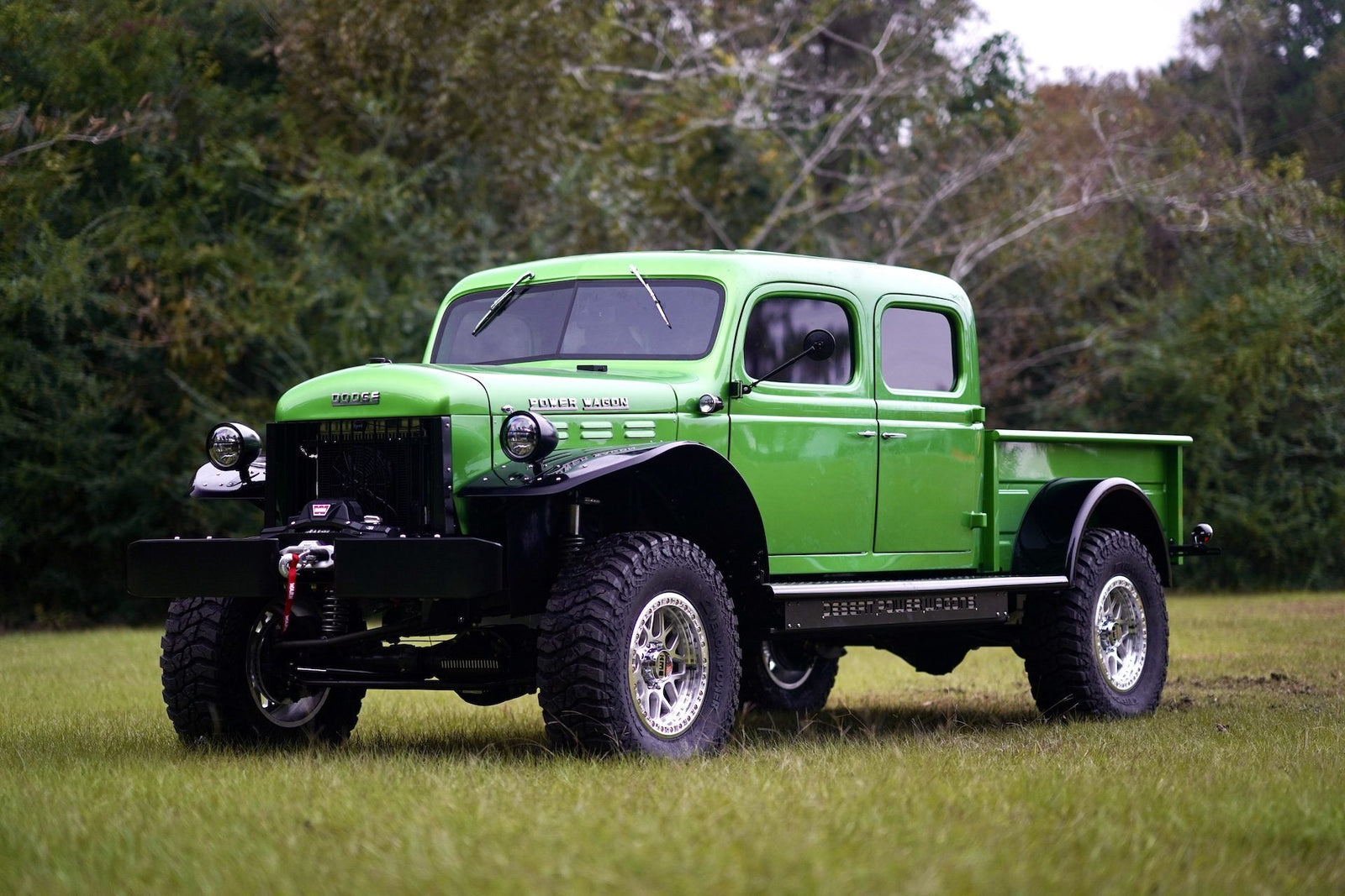 Bright green vintage Dodge Power Wagon truck with large off-road tires parked on grass, trees in the background.