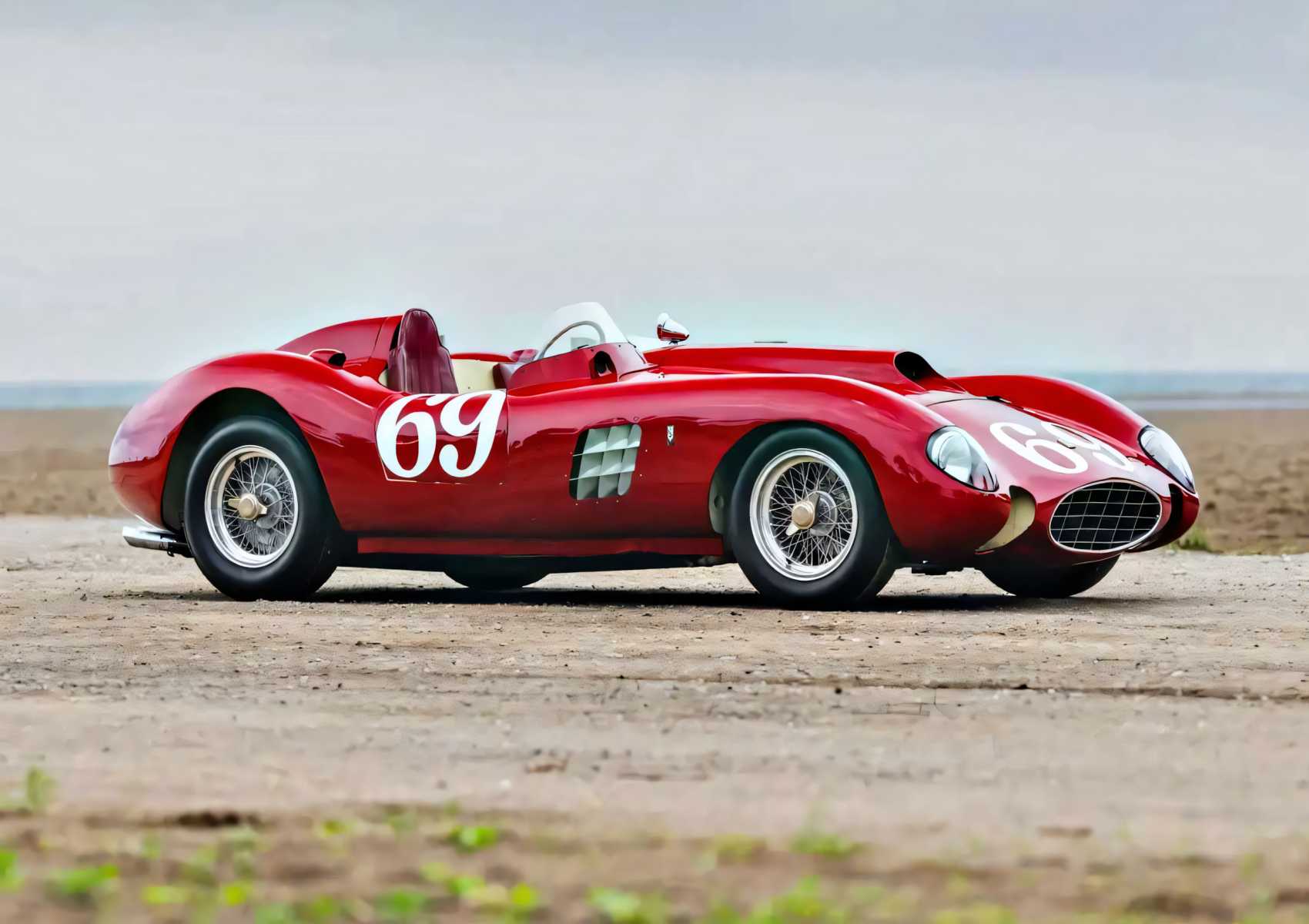 A classic red Ferrari 375 Plus race car with the number 69 on the side and hood is parked on a sandy surface under a cloudy sky.