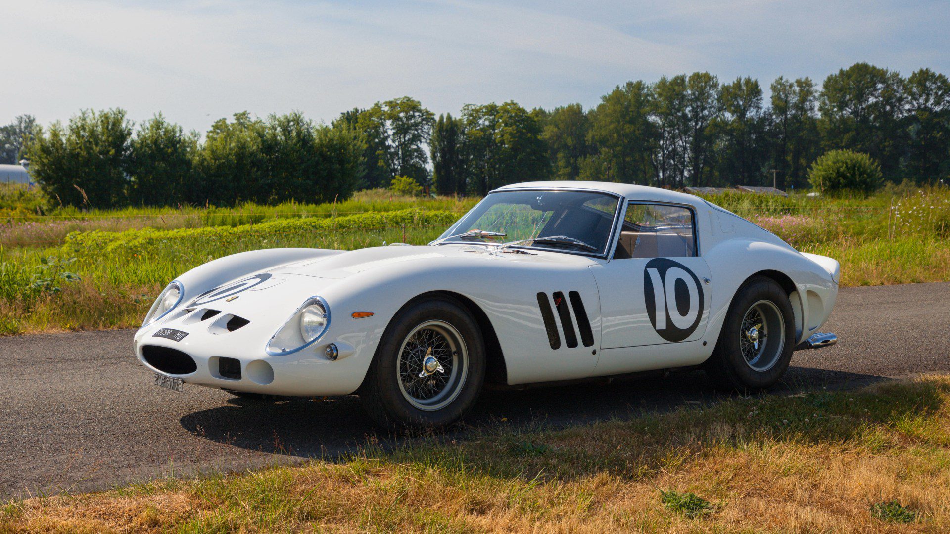 A factory white Ferrari 250 GTO with the number 10 on the door is parked on a rural road, surrounded by grass and trees under a clear sky—a stunning sight reminiscent of a Mecum Kissimmee Auction classic.